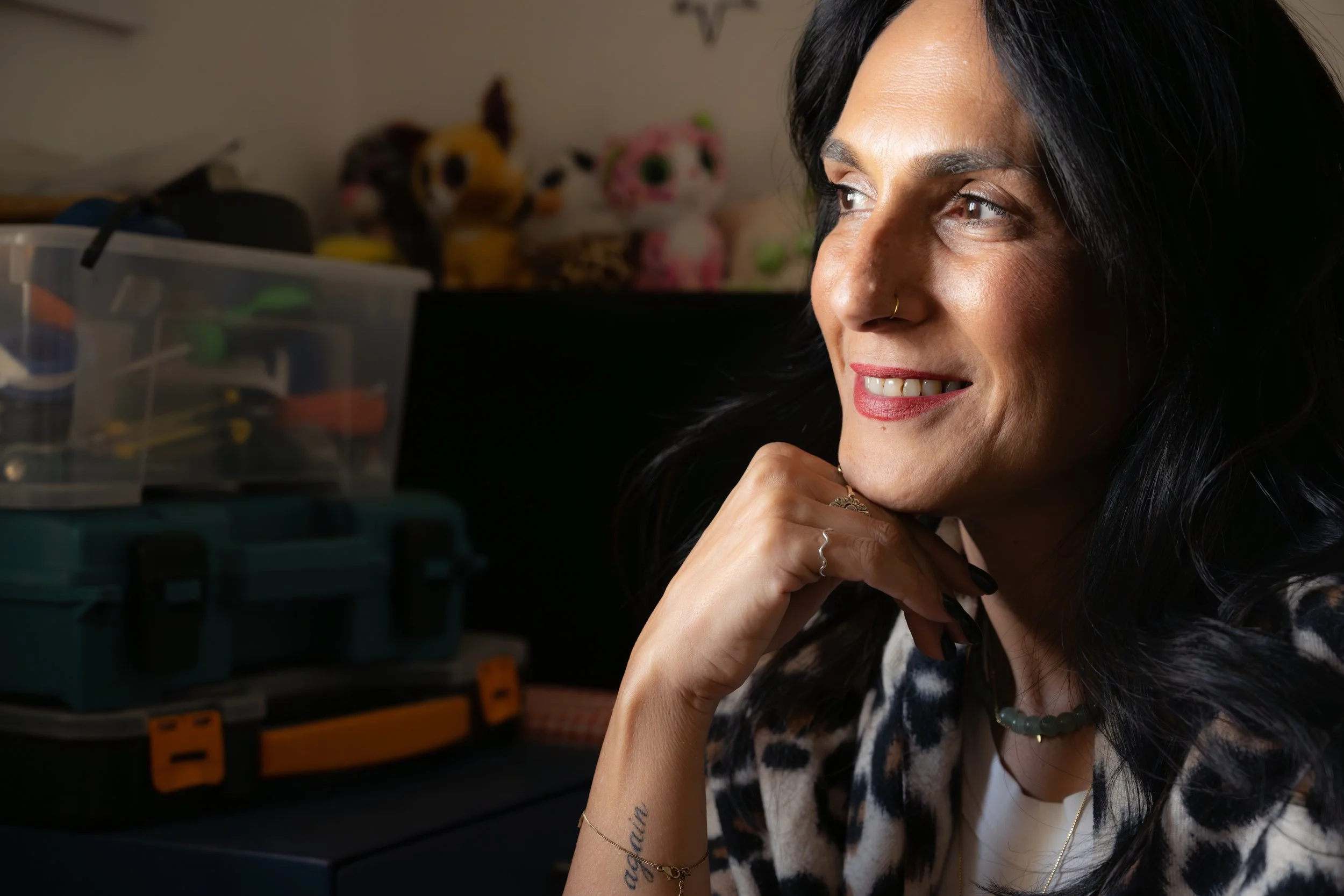 Limor gazes thoughtfully toward the light, smiling softly in a leopard-print jacket, with toolboxes and soft toys blurred in the background