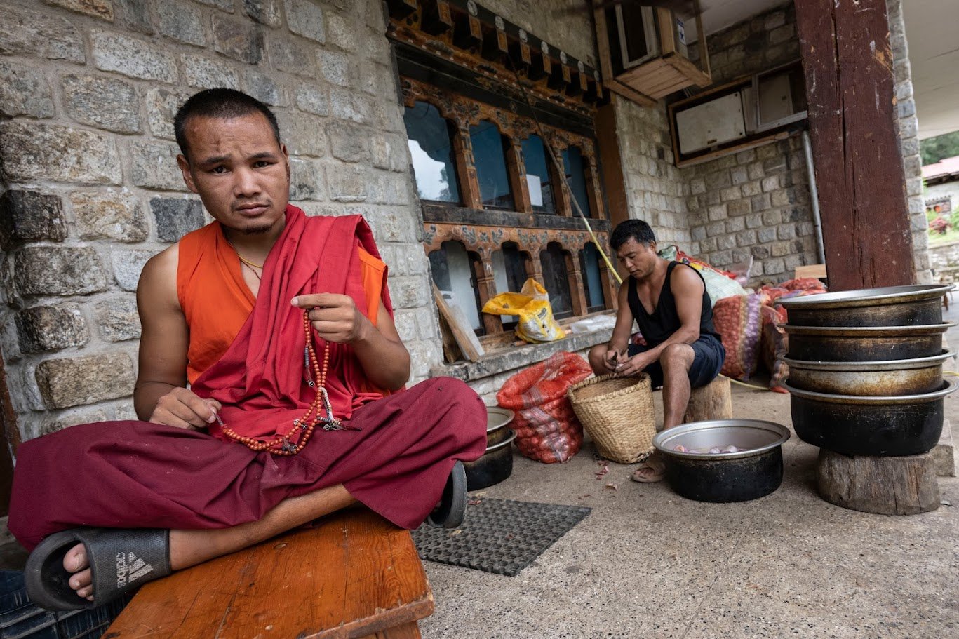 נזיר בודהיסטי יושב על מדרגה בכניסה לבית, מוקף בסירי מתכת מבריקים ובשכנים ברקע – בהוטן