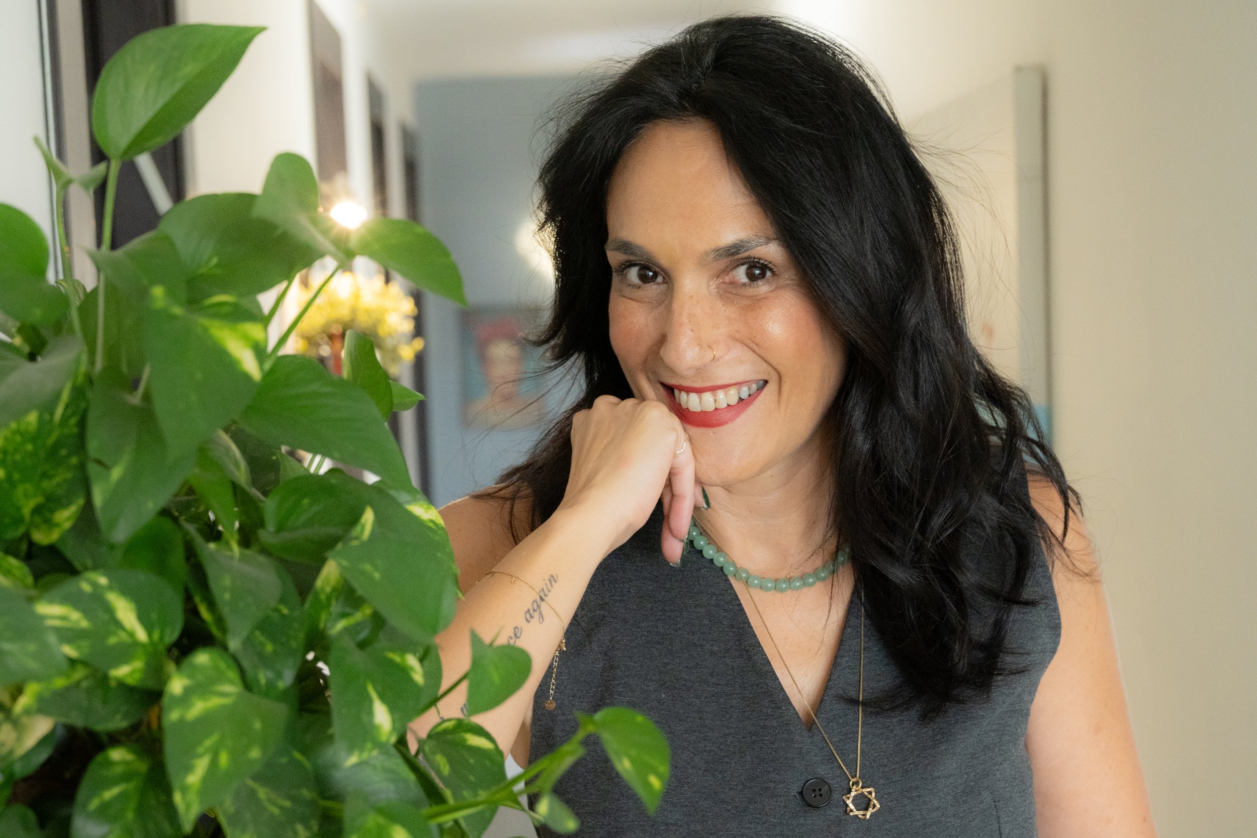 לימור פורן נשענת על מדף צהוב מלא בצמחייה ירוקה, מחייכת למצלמה במסדרון מואר, מוקפת צמחים ואור רך.