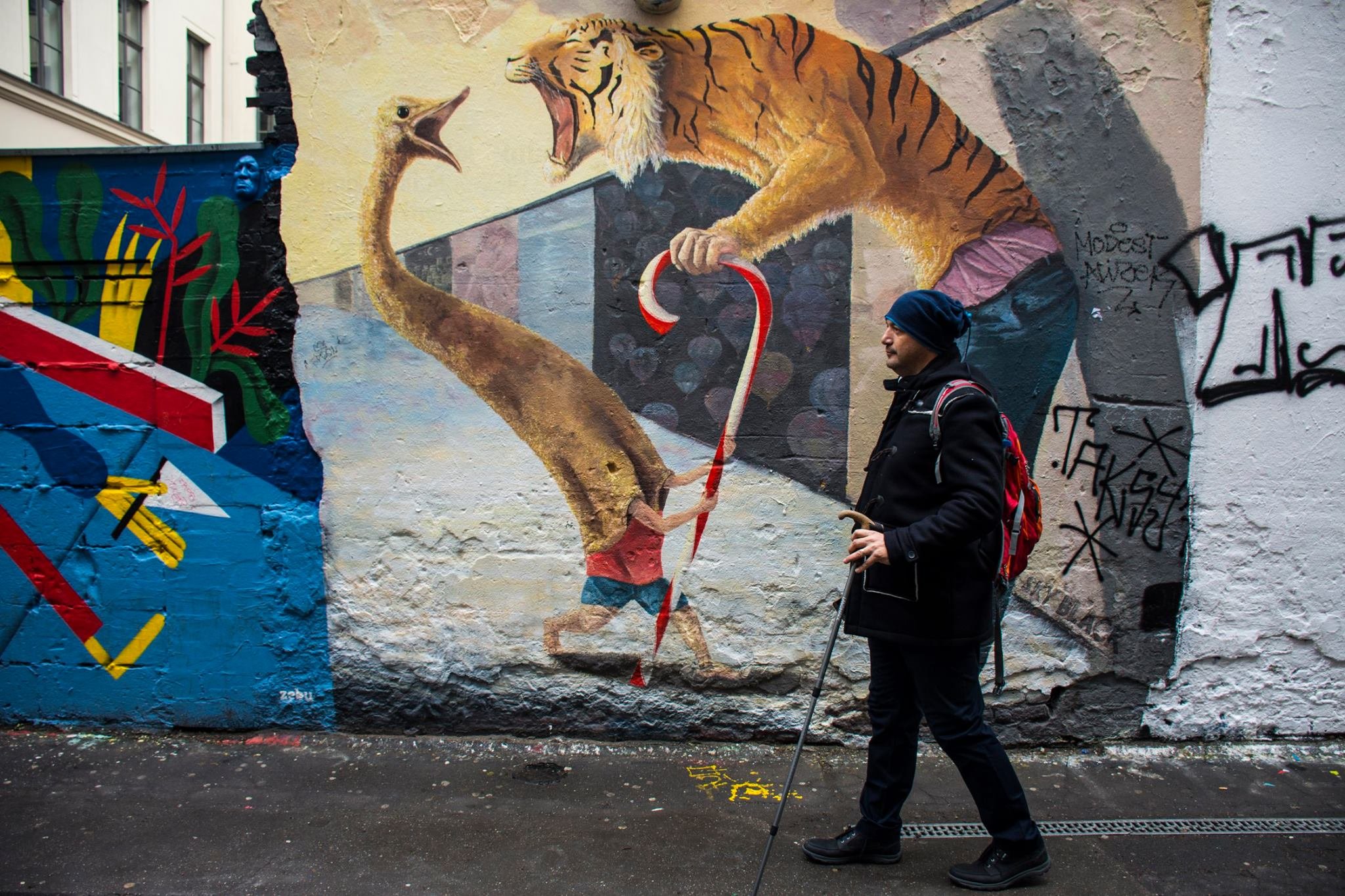 גבר עם מקל ותרמיל עובר ליד ציור קיר בסמטה בברלין של טיגריס ויען המחזיקים סוכריית מקל
