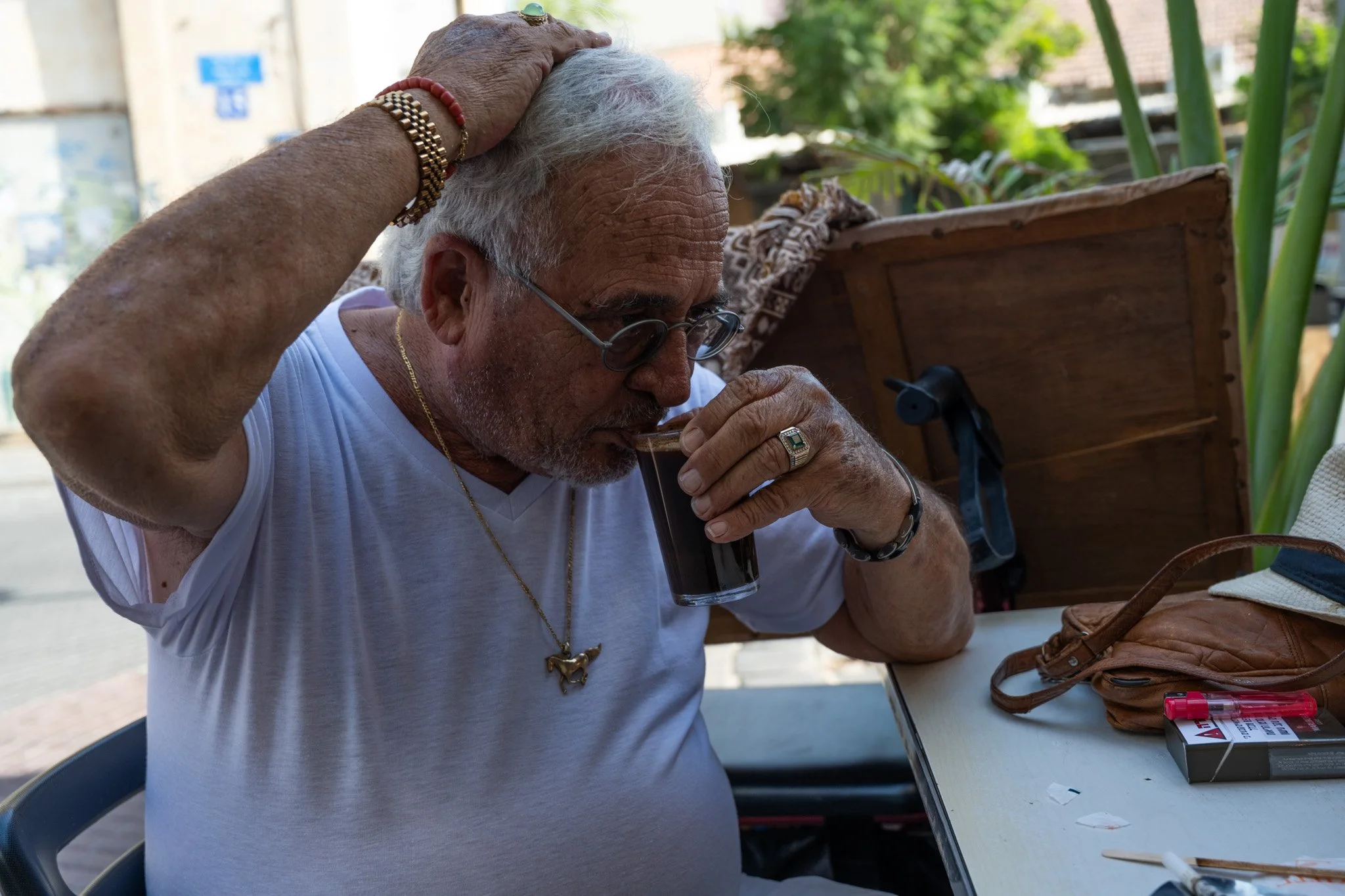 דוד דהן לבוש חולצה לבנה לוגם קפה חזק ליד שולחן בשוק הפשפשים, ידו על ראשו ותכשיטי זהב ומשקפיים נראים