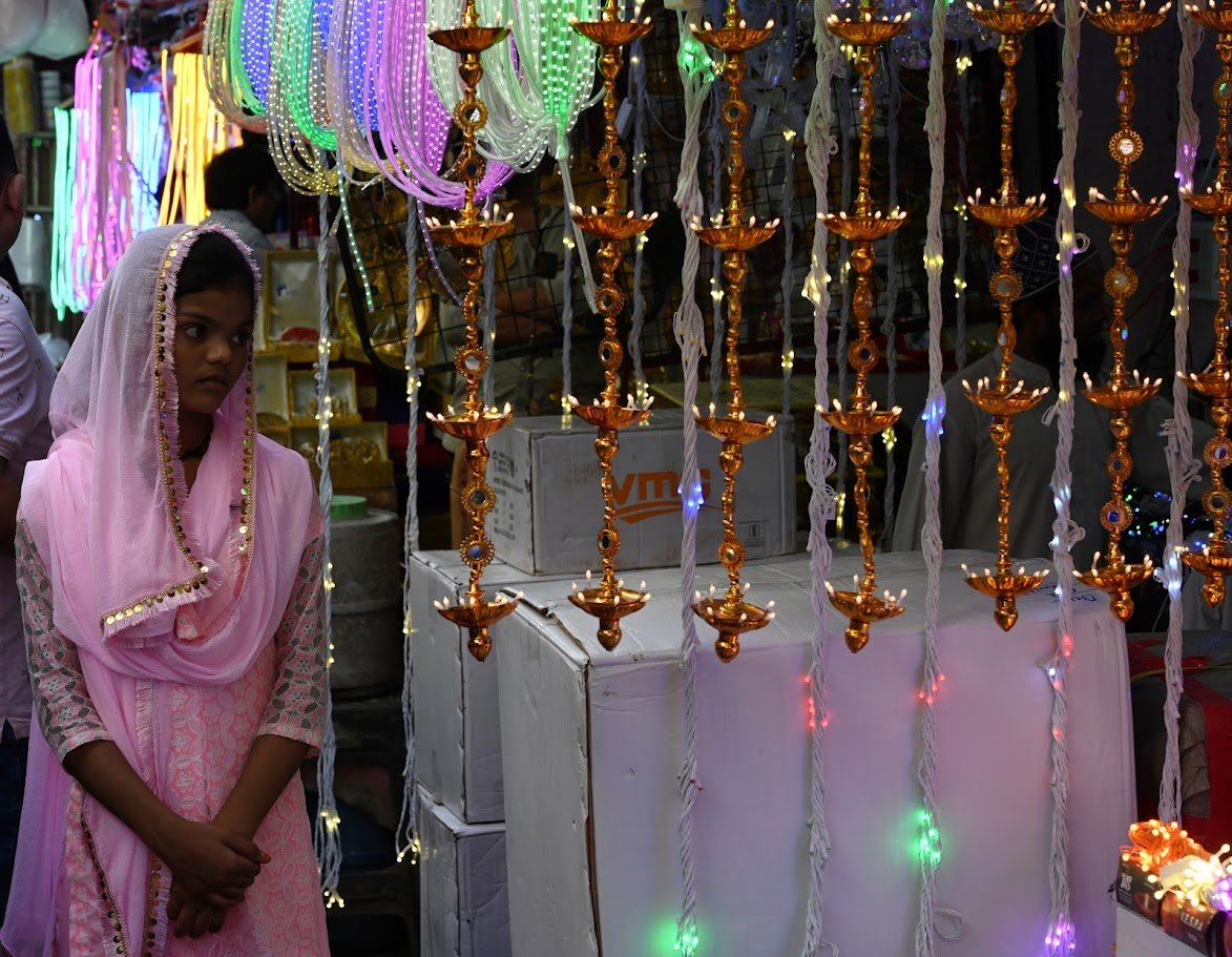 אישה צעירה בסארי ורוד עומדת בין שרשראות קישוטים צבעוניות ומנצנצות – דלהי, הודו
