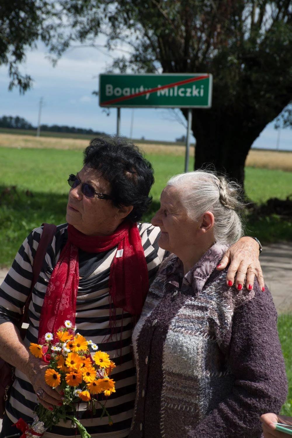 שתי נשים מבוגרות מתחבקות לצד הדרך ליד השלט של בוגוטי־מילצקי, אחת מחזיקה זר פרחים כתומים