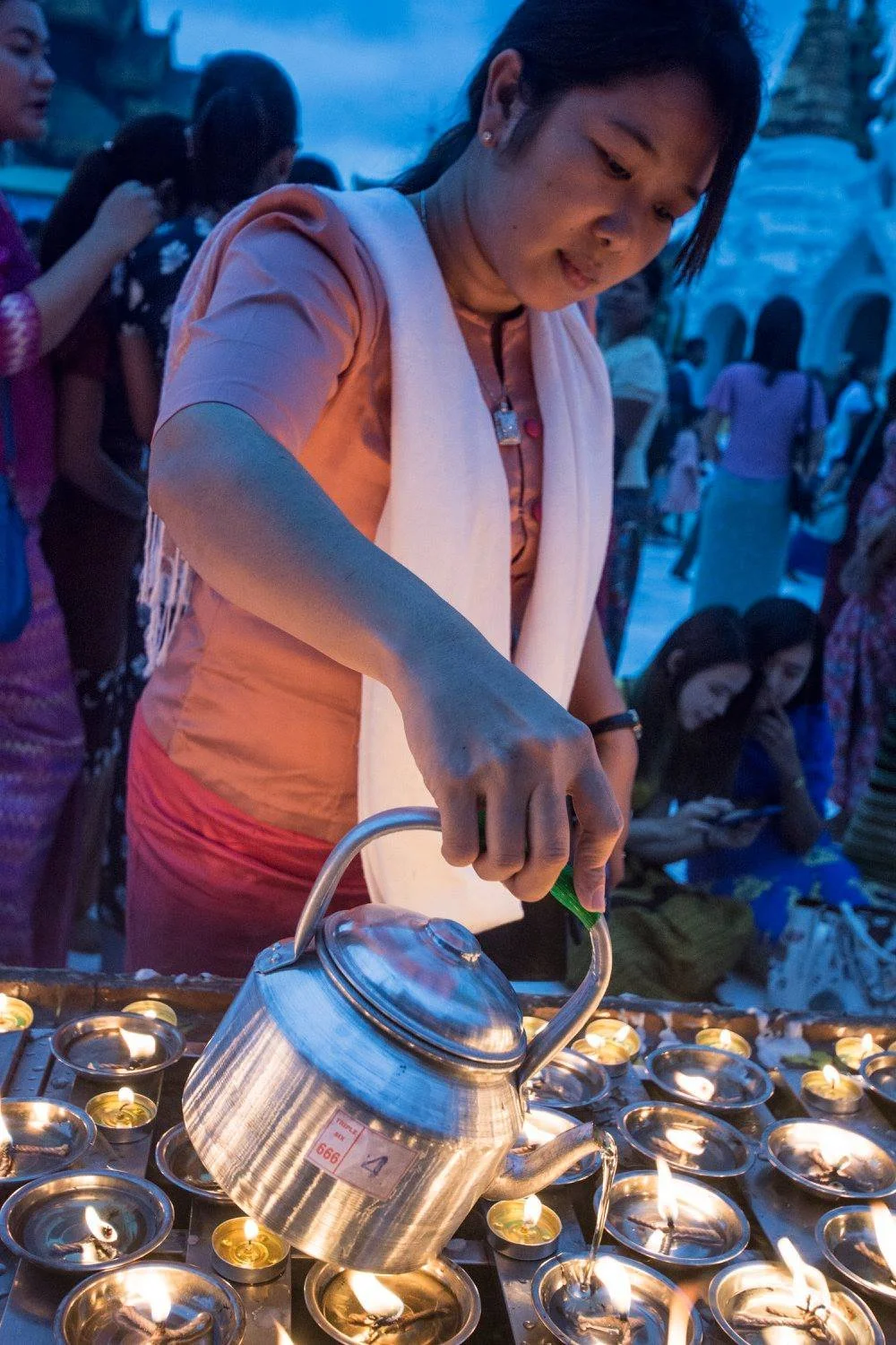 Woman pouring oil into small lamps during the Thadingyut Festival in Myanmar, surrounded by evening blue light and glowing flames
