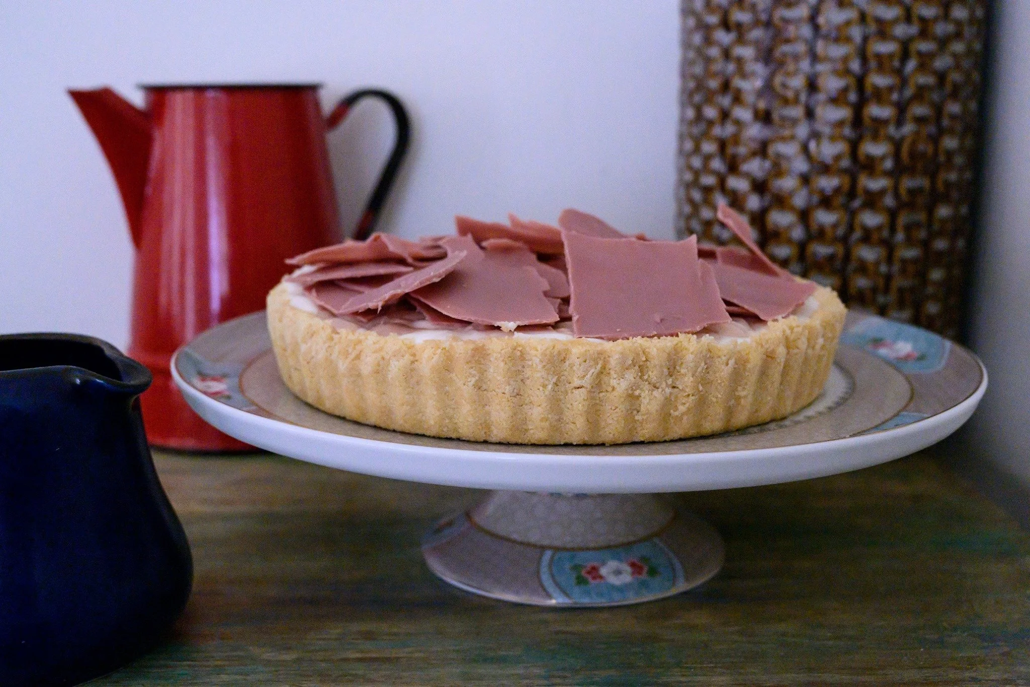 טארט שוקולד עם פתיתי שוקולד רובי מונח על סטנד וינטג' ומלווה בקנקן אמייל אדום — שקט של אחר הצהריים