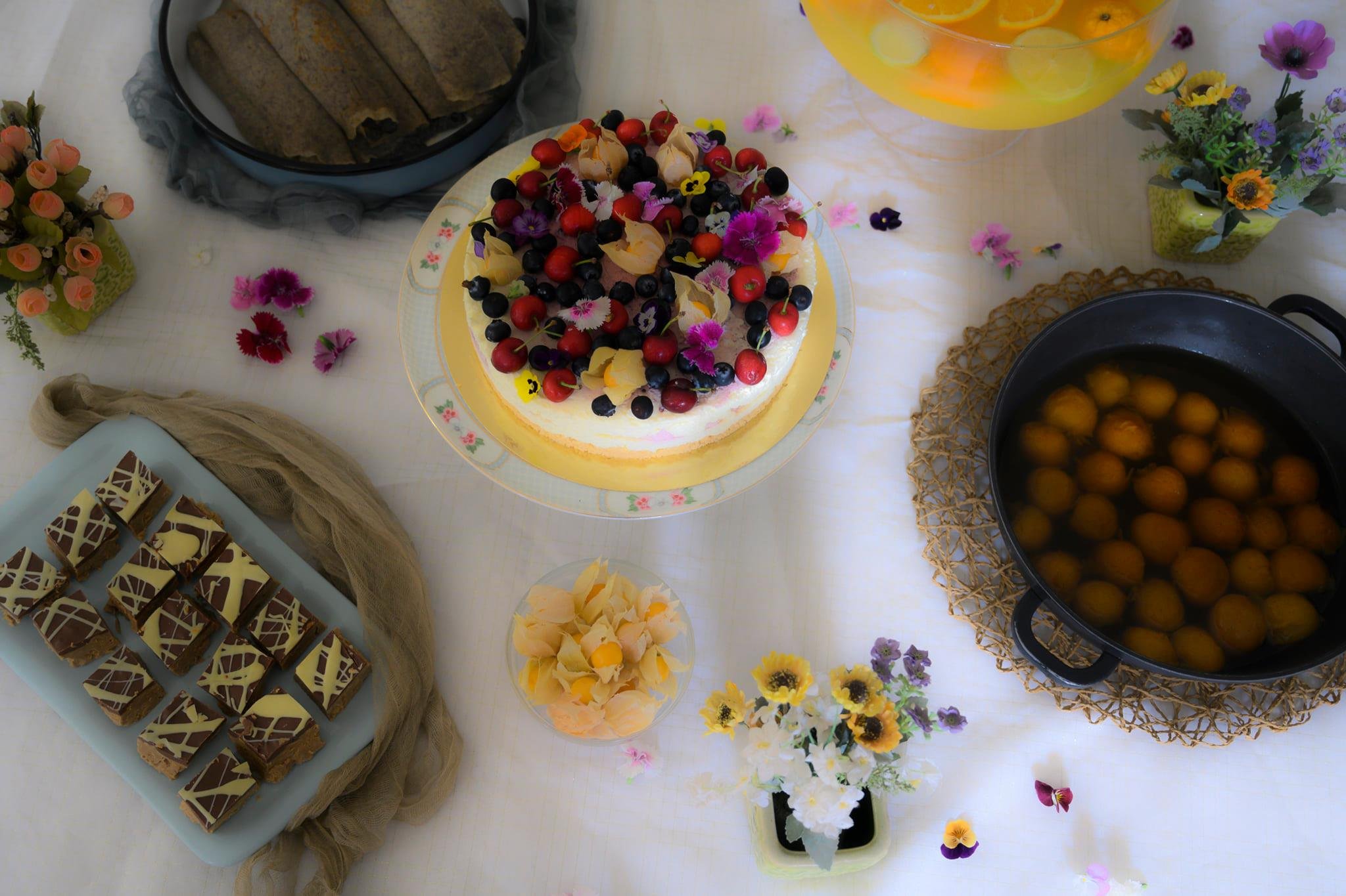 שולחן קינוחים אביבי מלא צבע: עוגת גבינה עם פירות יער במרכז, ריבועי שוקולד, כיסוני בצק ספוגי סירופ, בלינצ'ס תפוחים ואגוזים וקערות פרחים צבעוניות כמו המתוקים עצמם