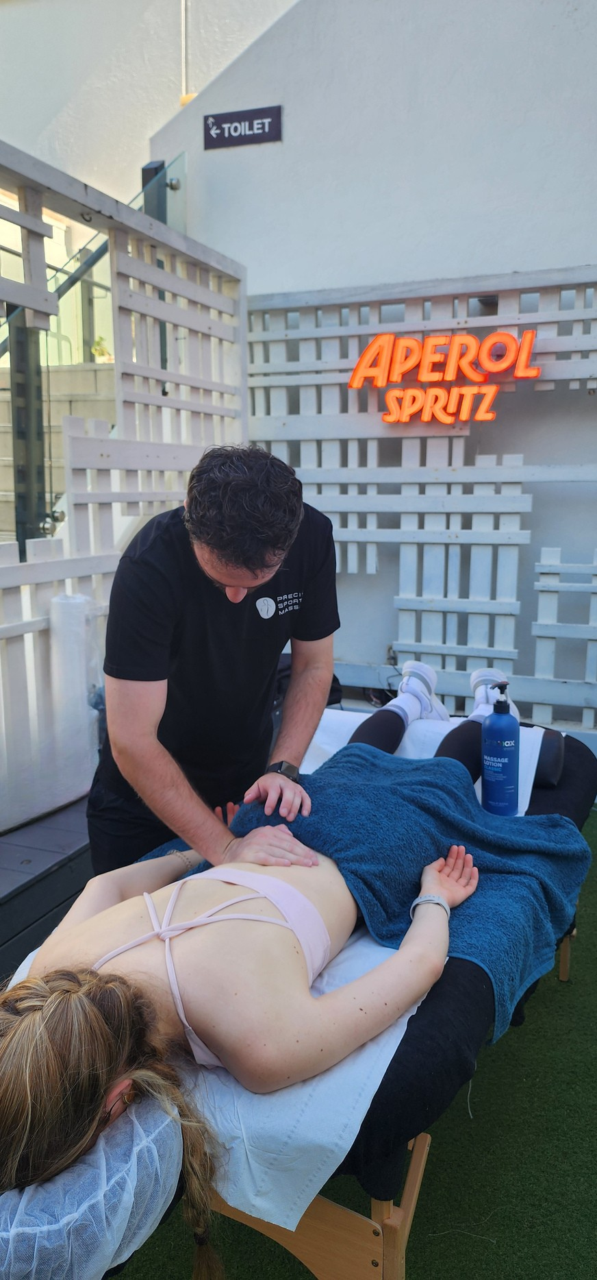 A person receiving a massage outdoors on a massage table, with a massage therapist working on their back, under a neon sign that says 'Aperol Spritz', with a sign for a toilet in the background.