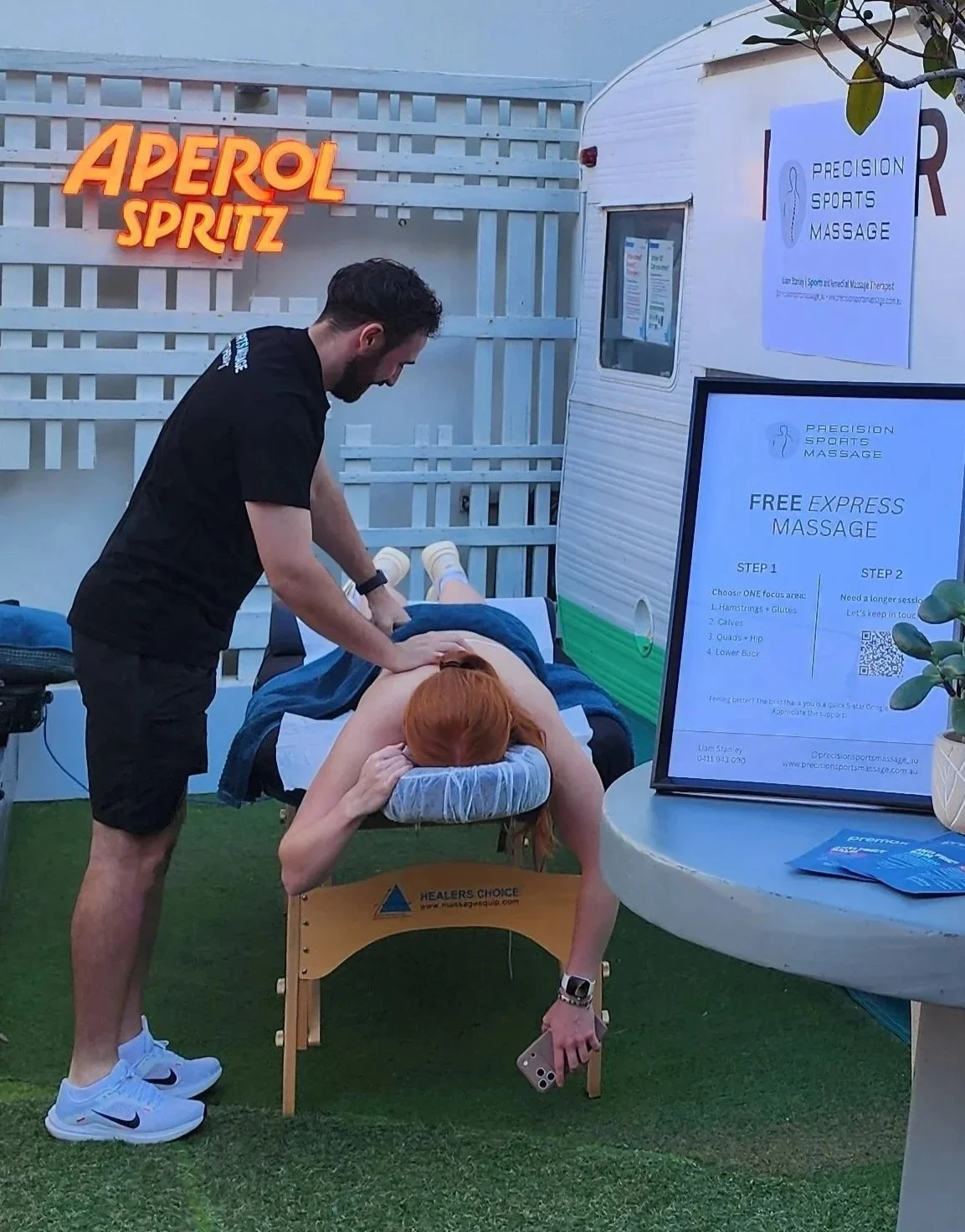 A massage therapist is giving a massage to a woman lying face down on a massage table outdoors, with a sign advertising free sports massage nearby.