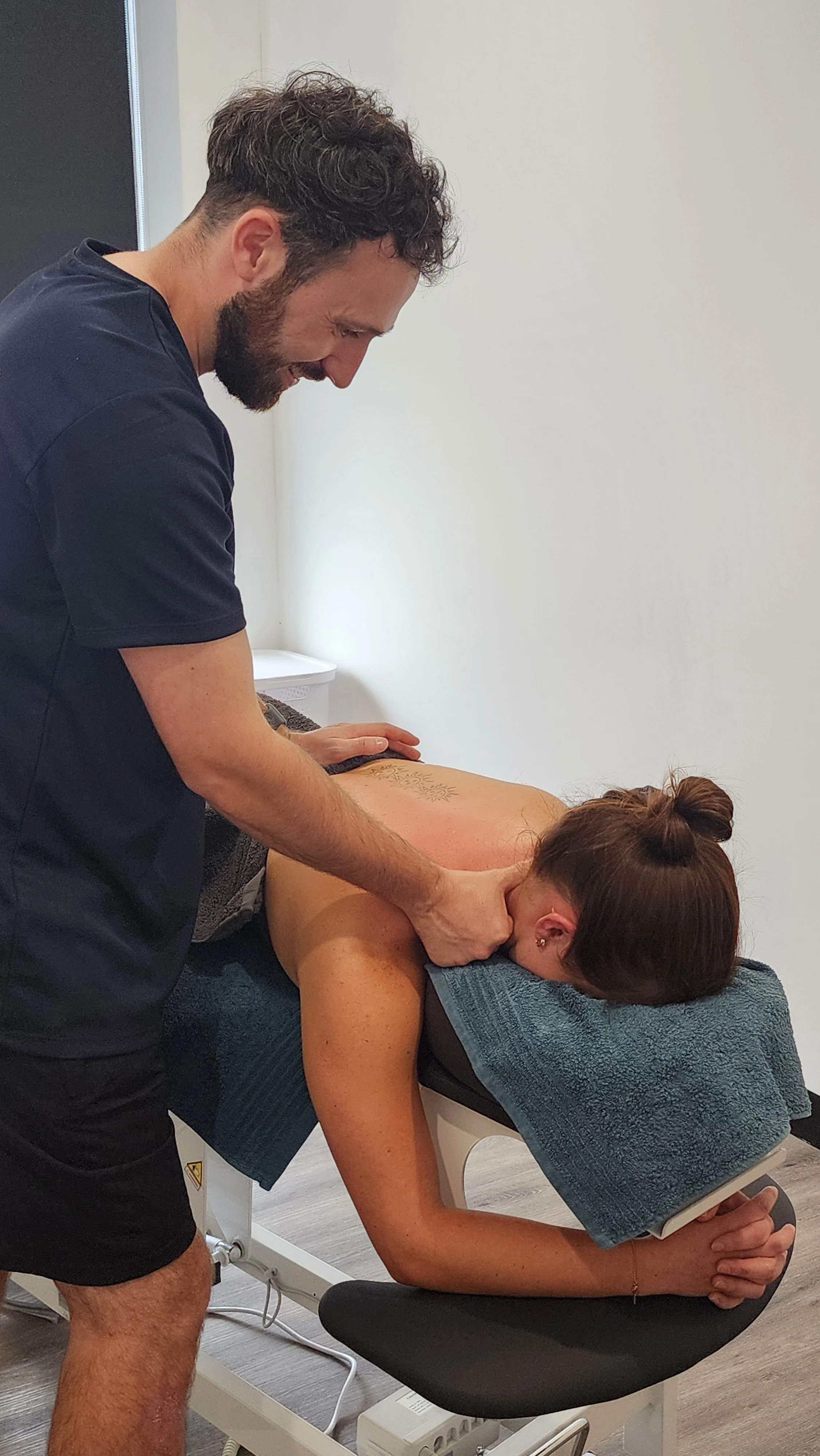 A person receiving a massage in a massage therapy session, laying face down on a massage table with a towel over their lower back, while a massage therapist stands beside them applying pressure to their neck and shoulder area.