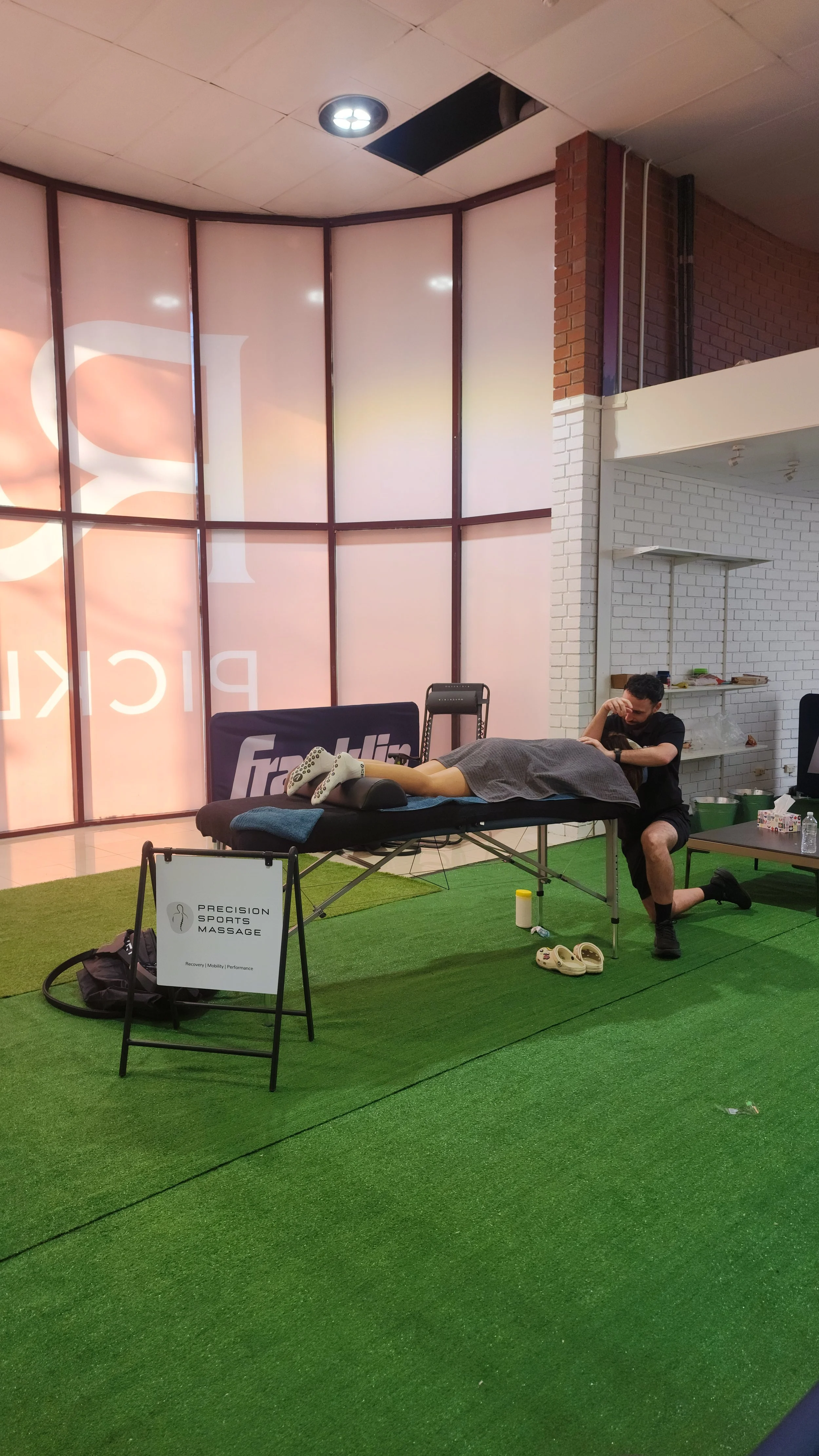 A man kneeling on green turf receiving a massage from a woman lying face down on a massage table, with a sign for a sports massage service in the foreground and a store with pink and white walls in the background.