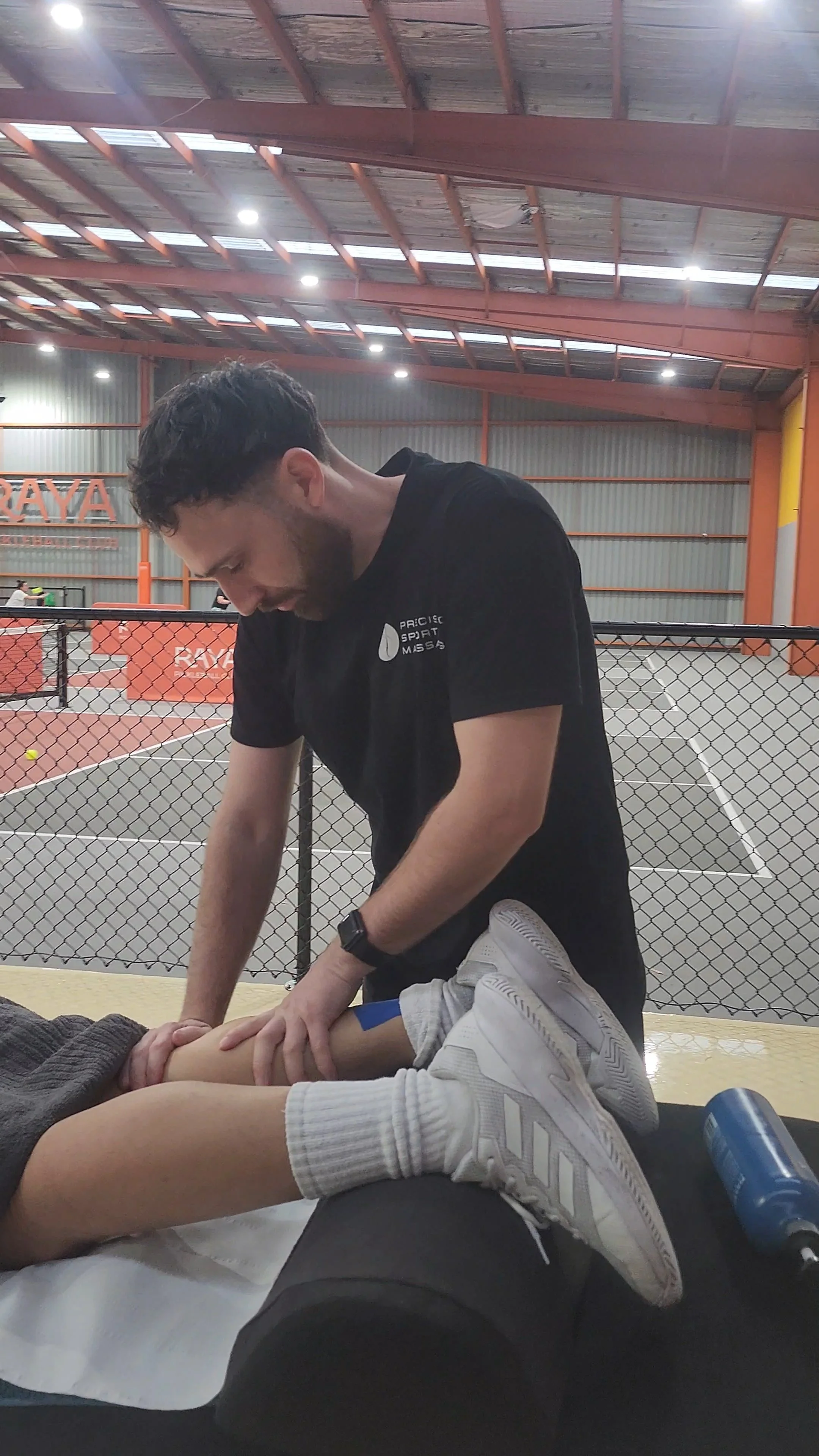 A person in sportswear lying on the ground receiving first aid from a man in a black shirt inside an indoor sports facility with a tennis or pickleball court in the background.