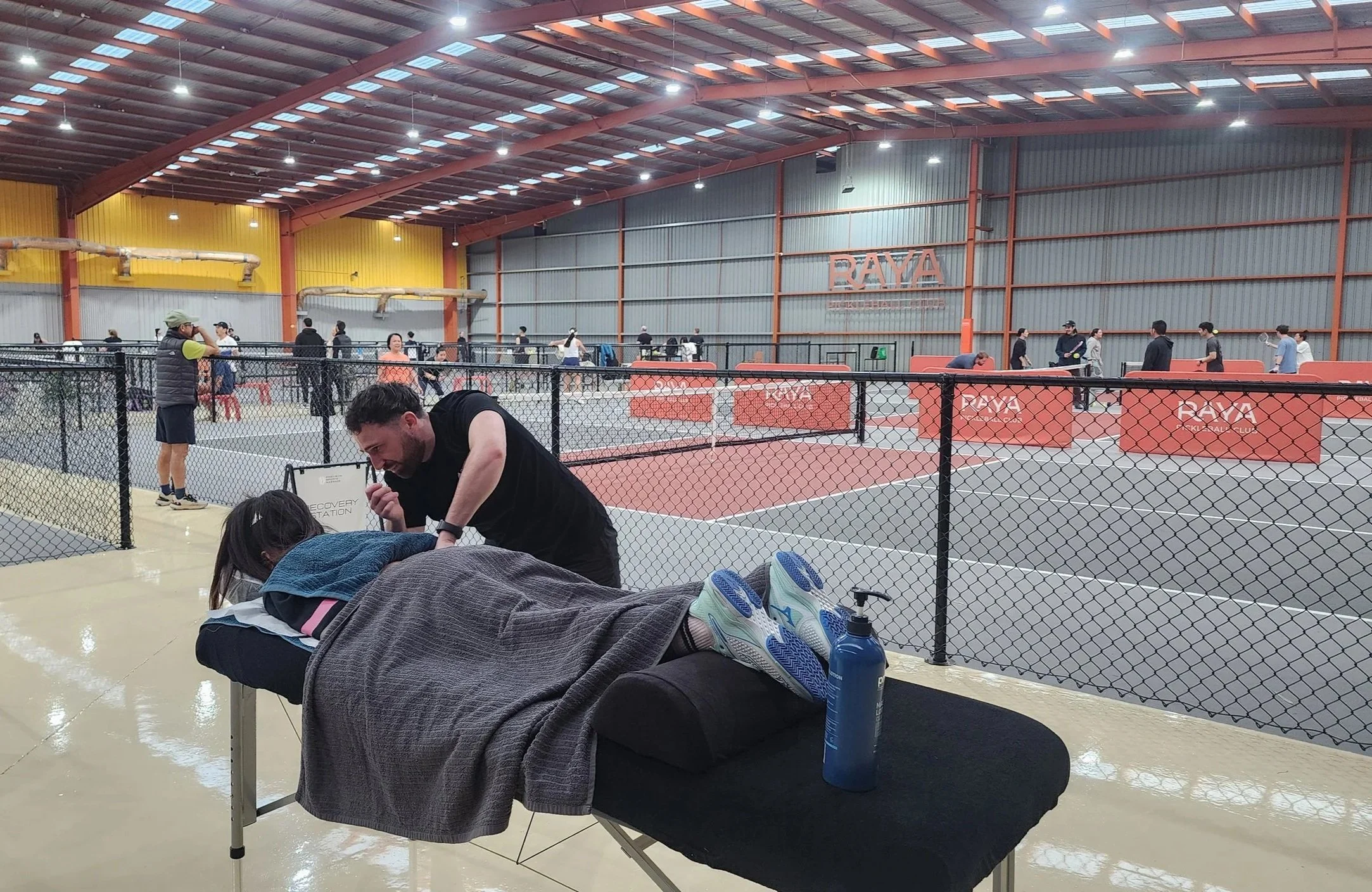 Indoor tennis or pickleball court with people playing, sitting, and walking, separated by a black chain-link fence, with large yellow, gray, and red walls, and a high metal roof with skylights.