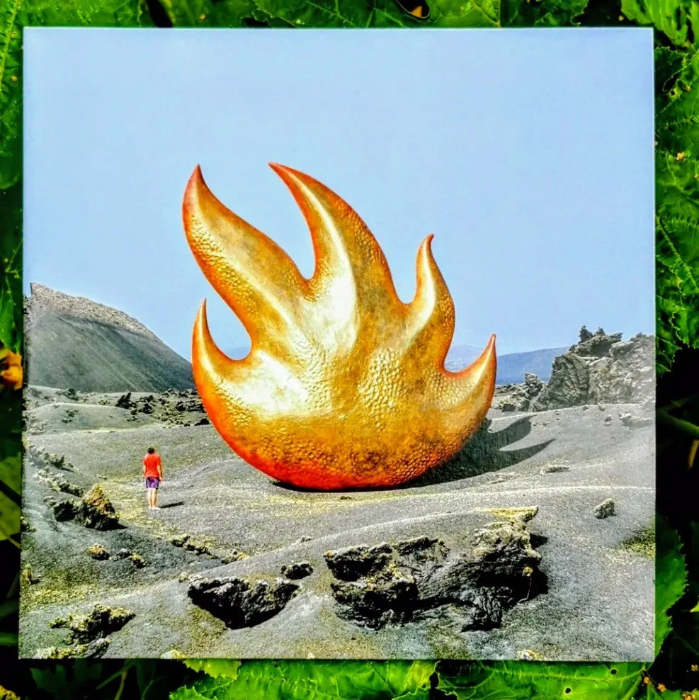 A person standing in a rocky, barren landscape with a large, fiery-shaped sculpture resembling a flame nearby. The sky is clear and blue.