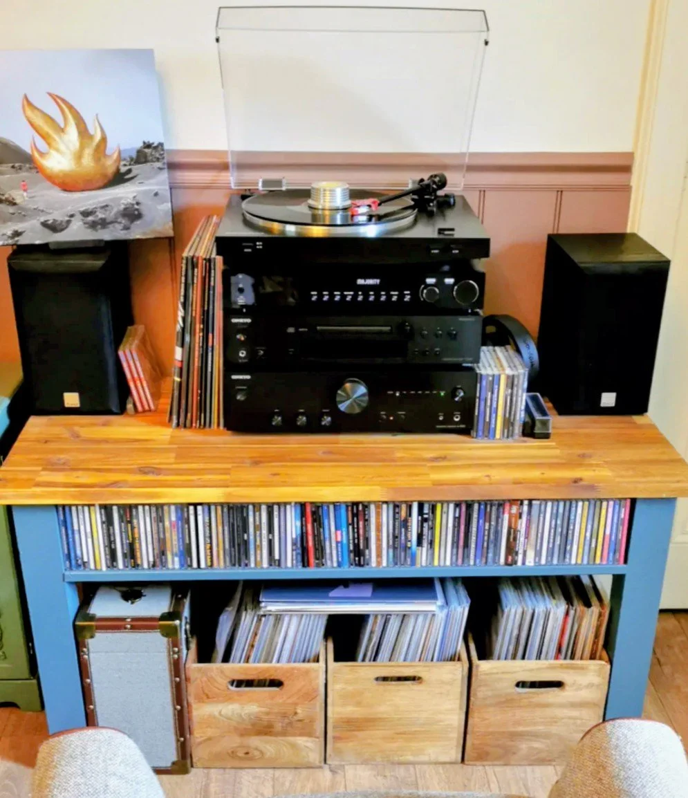 A vintage stereo system on a wooden table with a turntable, speakers, vinyl records, and shelves filled with records and storage bins.