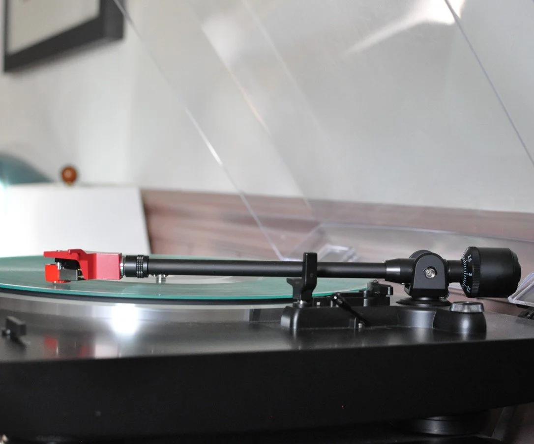 Close-up of a turntable playing a vinyl record, with a black tonearm and red cartridge.