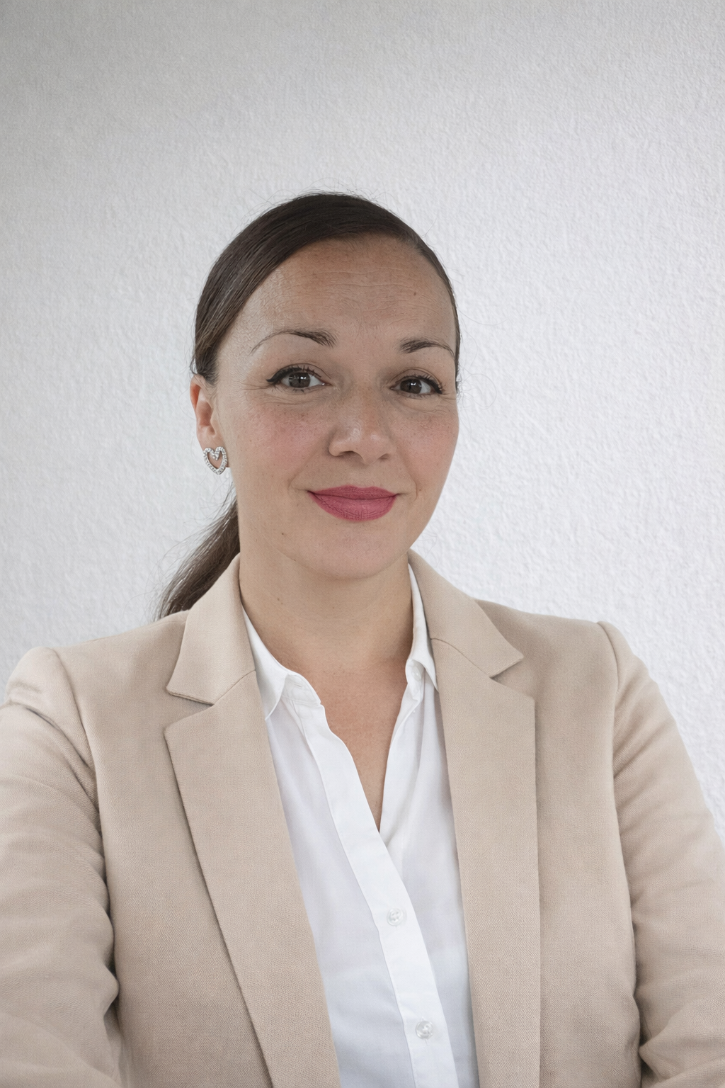 Ein Porträt einer Frau mit braunen Haaren, die einen cremefarbenen Blazer und weiße Bluse trägt, vor einer weißen Wand.