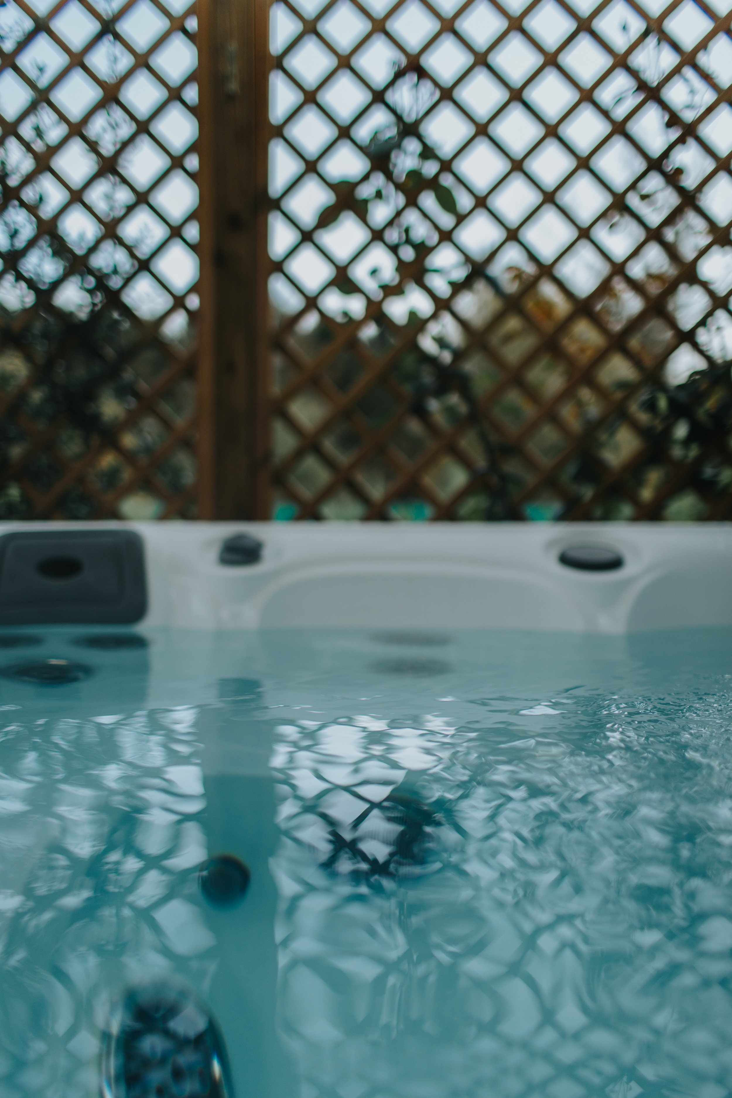 A hot tub filled with water, with a wooden lattice fence and some greenery in the background.