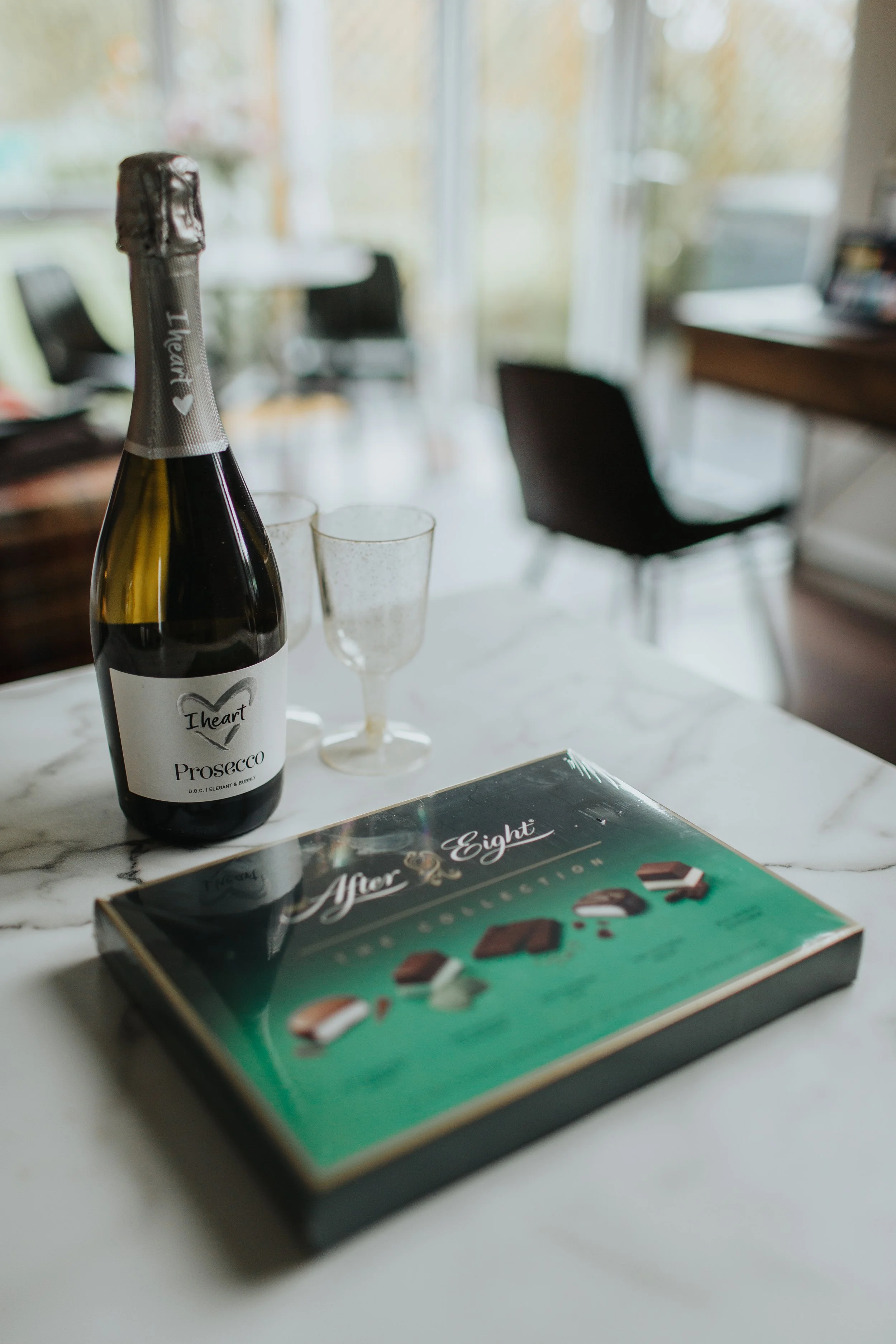 A bottle of sparkling wine labeled "Prosecco" next to two empty glasses and a box of chocolates on a white marble table in a bright room with large windows and blurred furniture in the background.