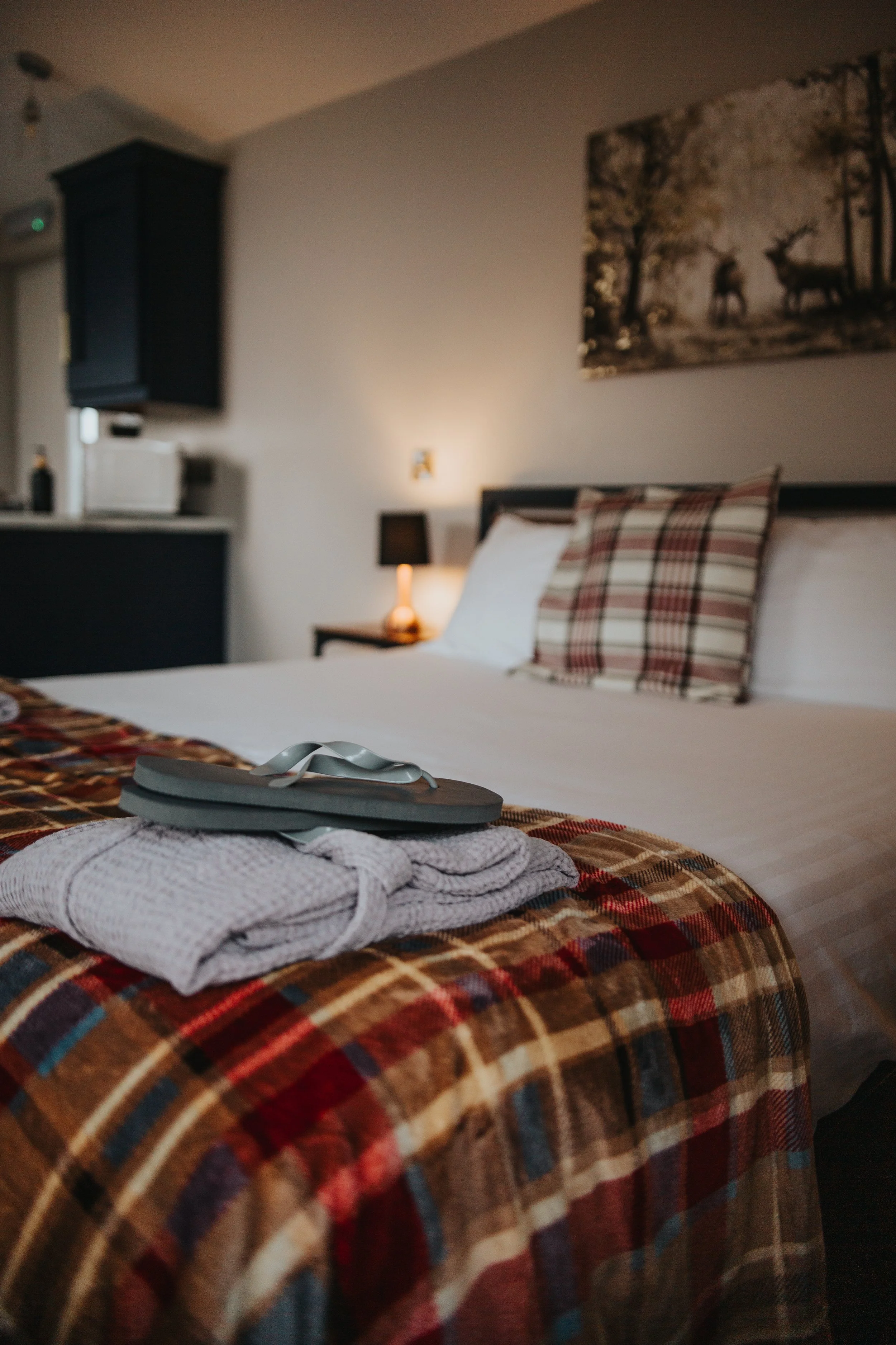 A neatly made bed with a checkered pillow and a folded towel and slippers on a colorful plaid bedspread in a cozy bedroom.