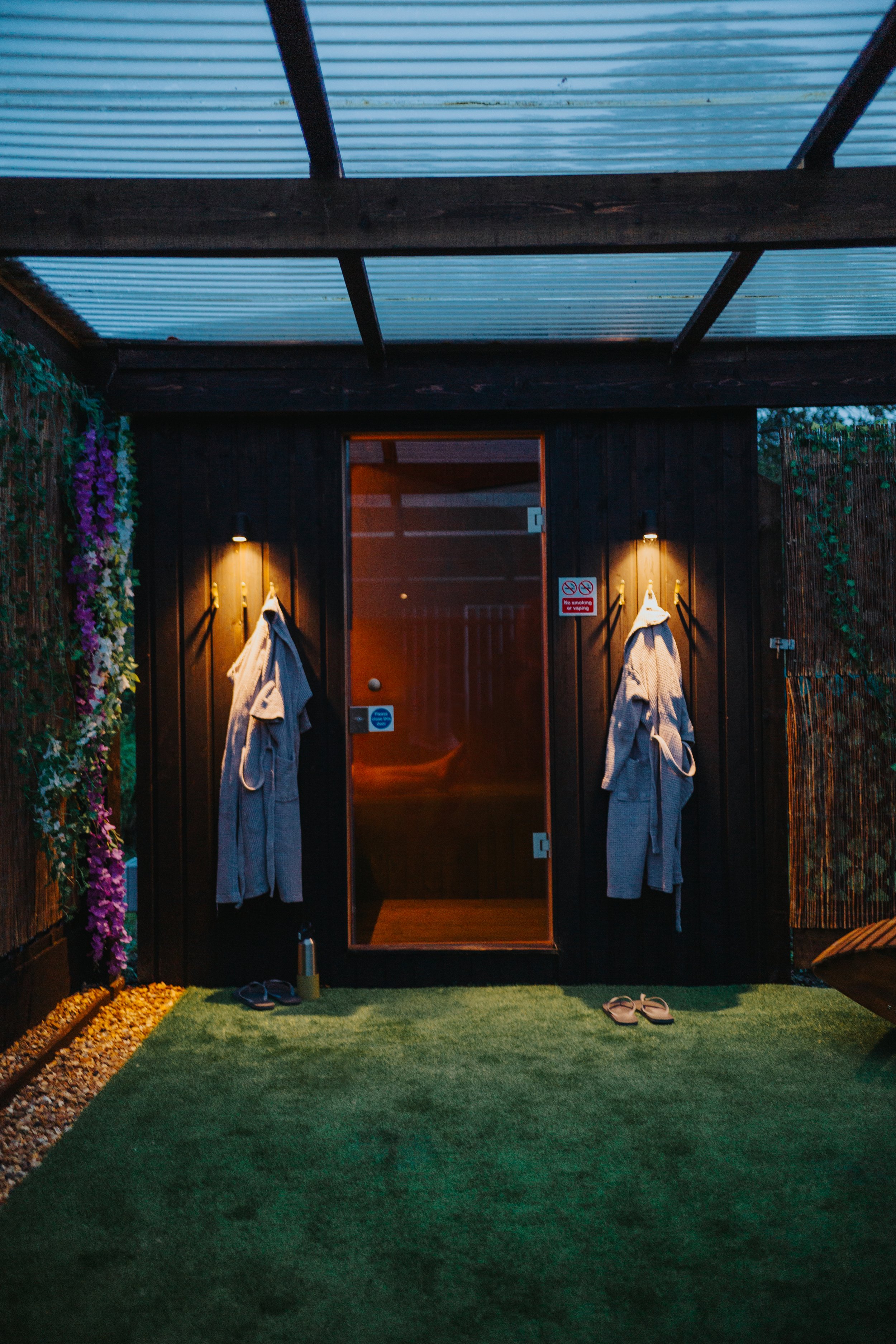 Outdoor spa or sauna area with a wooden door, two robes hanging on black hooks, soft lighting, a pair of slippers, and a drinking bottle on a green artificial grass mat, with decorative flowers and foliage on the sides.