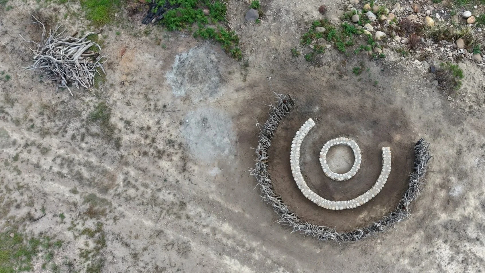 An aerial view of a dry, open landscape with patches of green vegetation. In the center, there is a spiral pattern made of white stones, surrounded by a ring of intertwined sticks or driftwood.