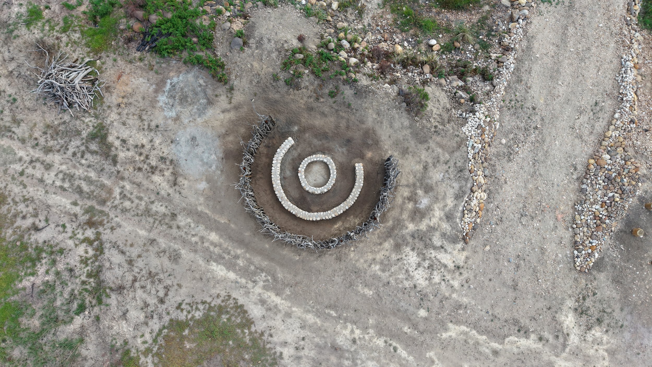 A spiral design made of white rocks in a circular outline on dirt ground, surrounded by natural landscape including rocks, sparse vegetation, and a pile of driftwood nearby.