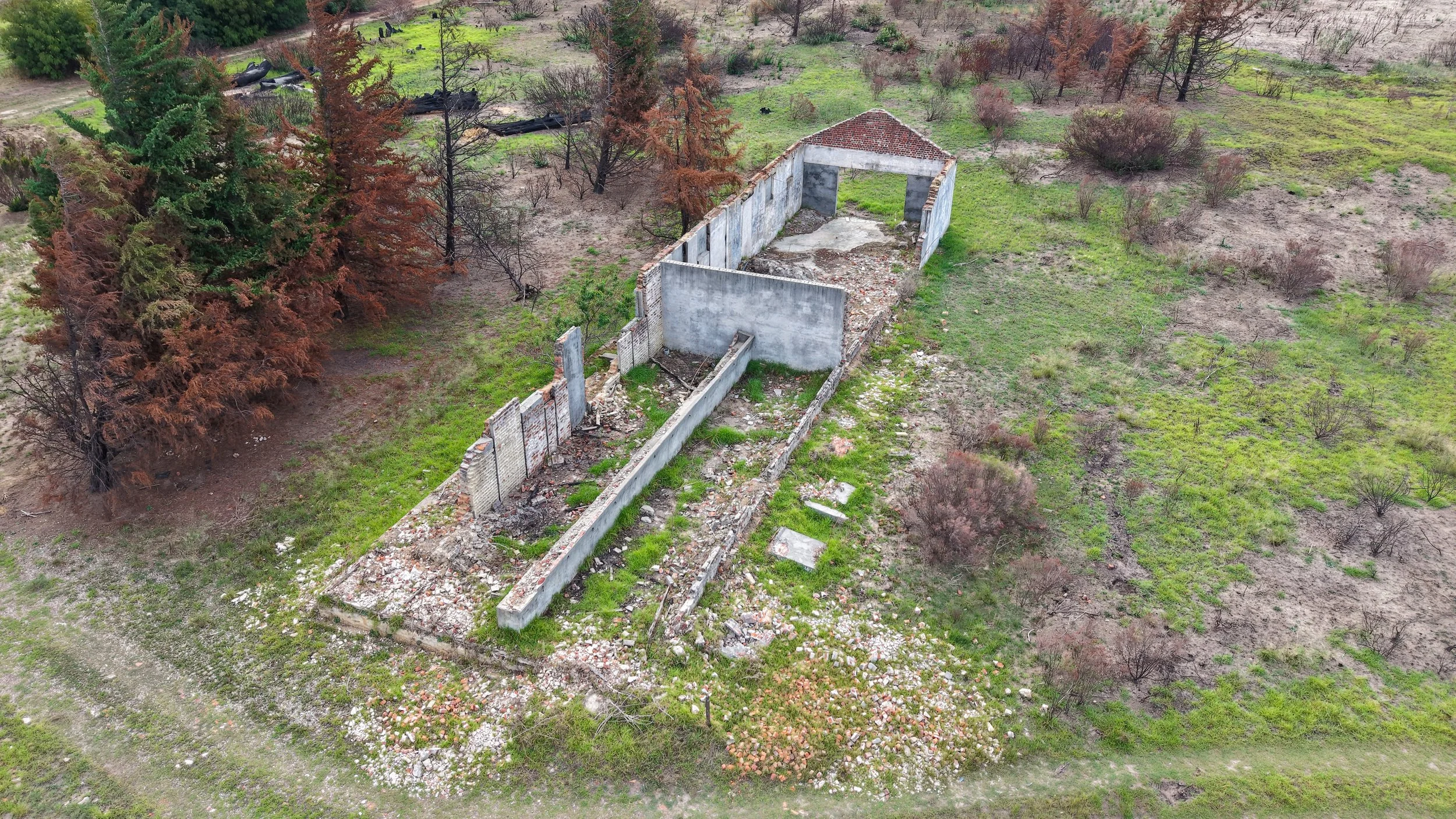 Aerial view of a destroyed building with back walls intact, surrounded by green grass, scattered debris, and leafless trees, with some coniferous trees nearby.