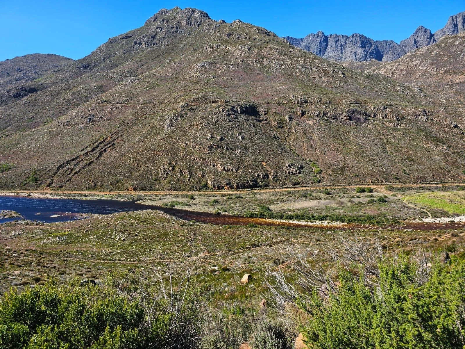 A mountainous landscape with a large steep hill or mountain covered in sparse vegetation. At the base, there is a river flowing, with a dirt road running parallel to it. The sky is clear and blue.