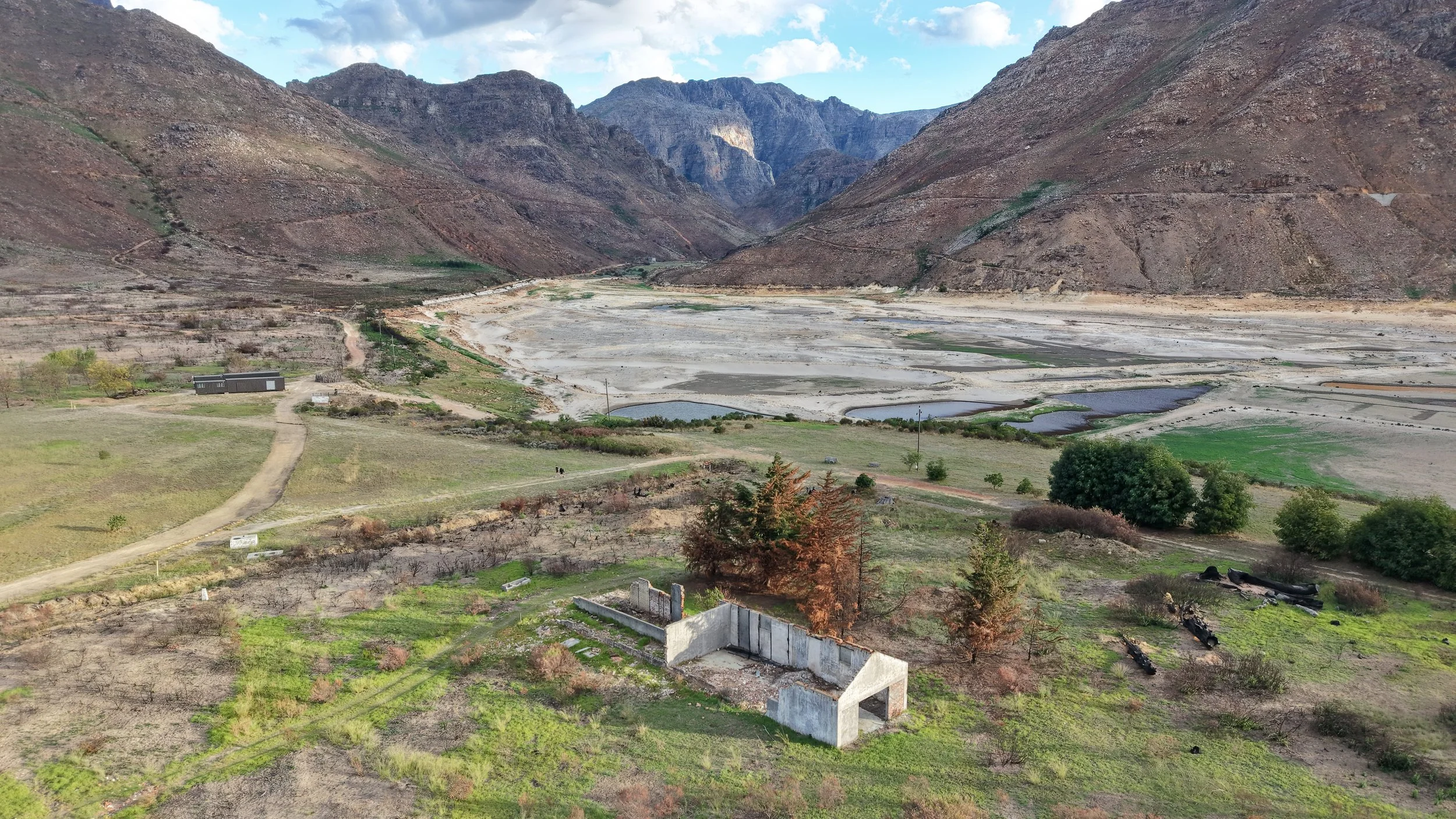 A rugged mountain landscape with sparse vegetation, a dry riverbed, and a few small ponds in a valley. There are some buildings, trees, and a dirt path in the foreground.