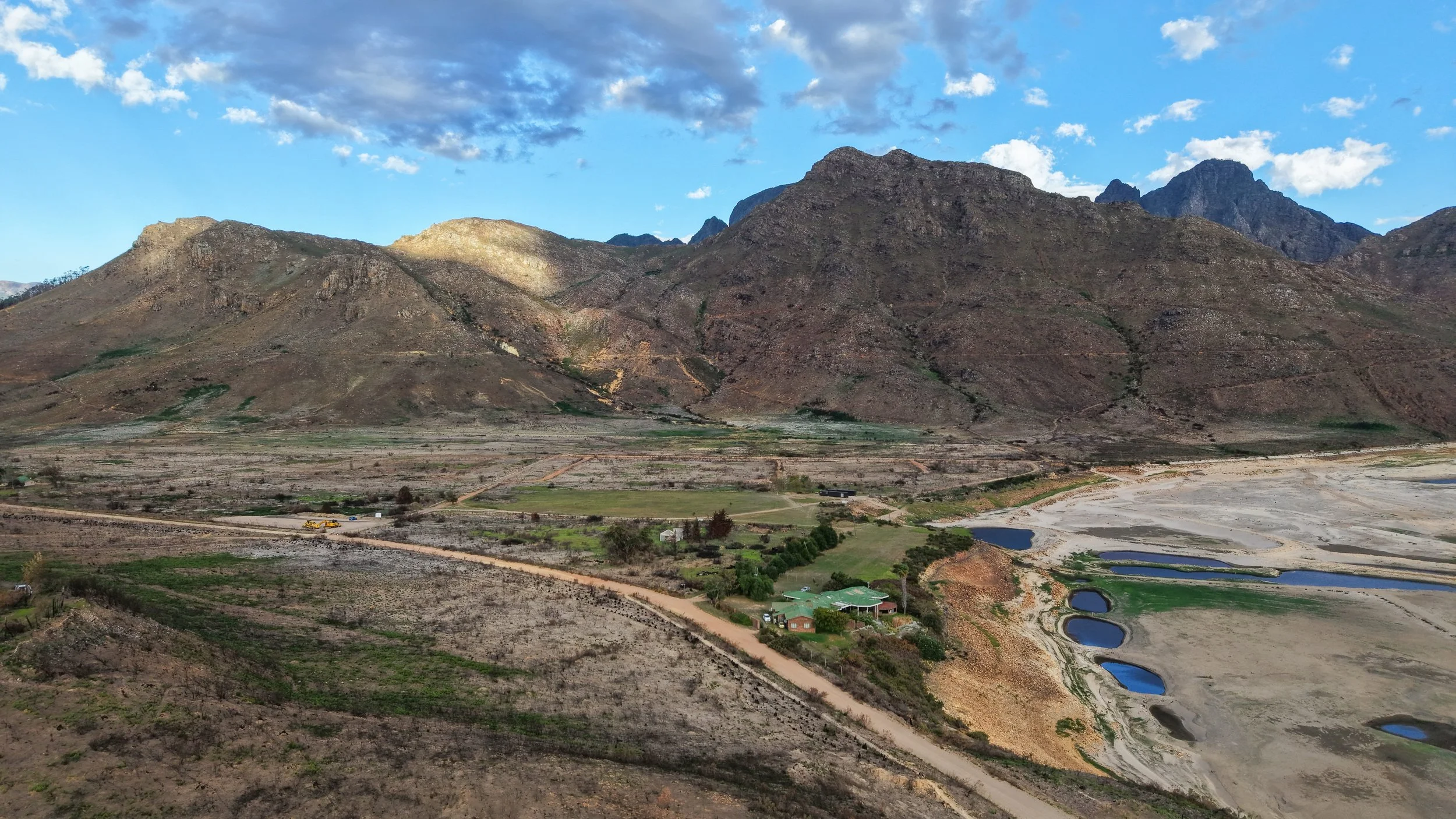 Scenic view of mountain range with a mix of rocky and forested areas under a blue sky with scattered clouds, and a dry riverbed with small ponds and a few buildings in the foreground.