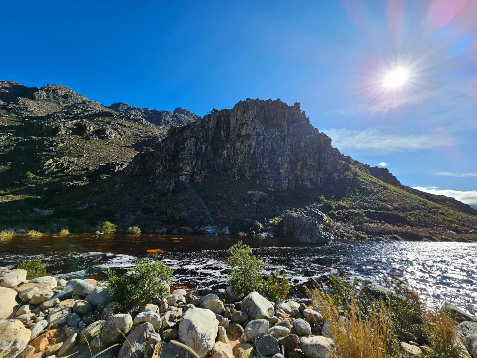 A landscape of rocky mountains with a river at the base, greenery and small bushes in the foreground, and a bright sun in a clear blue sky.