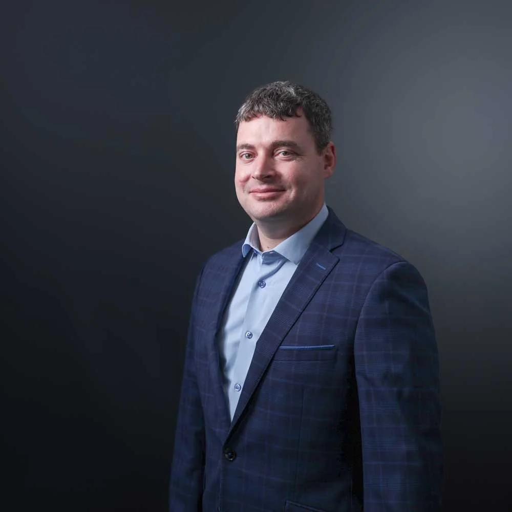 A professional man in a dark blue plaid suit and light blue shirt standing against a dark background.