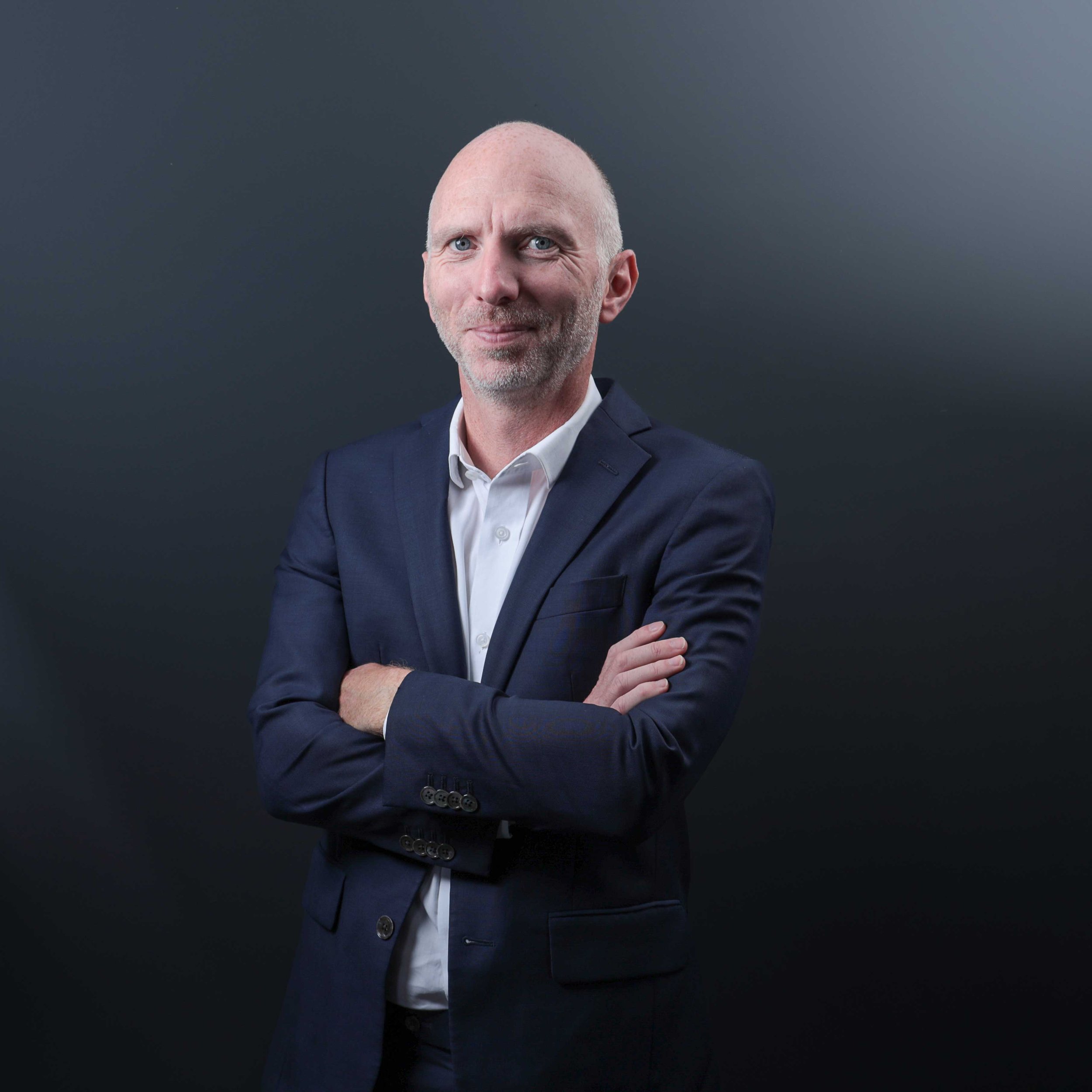 A middle-aged man with a bald head and gray beard, wearing a navy blue suit and white shirt, standing with arms crossed against a dark background.