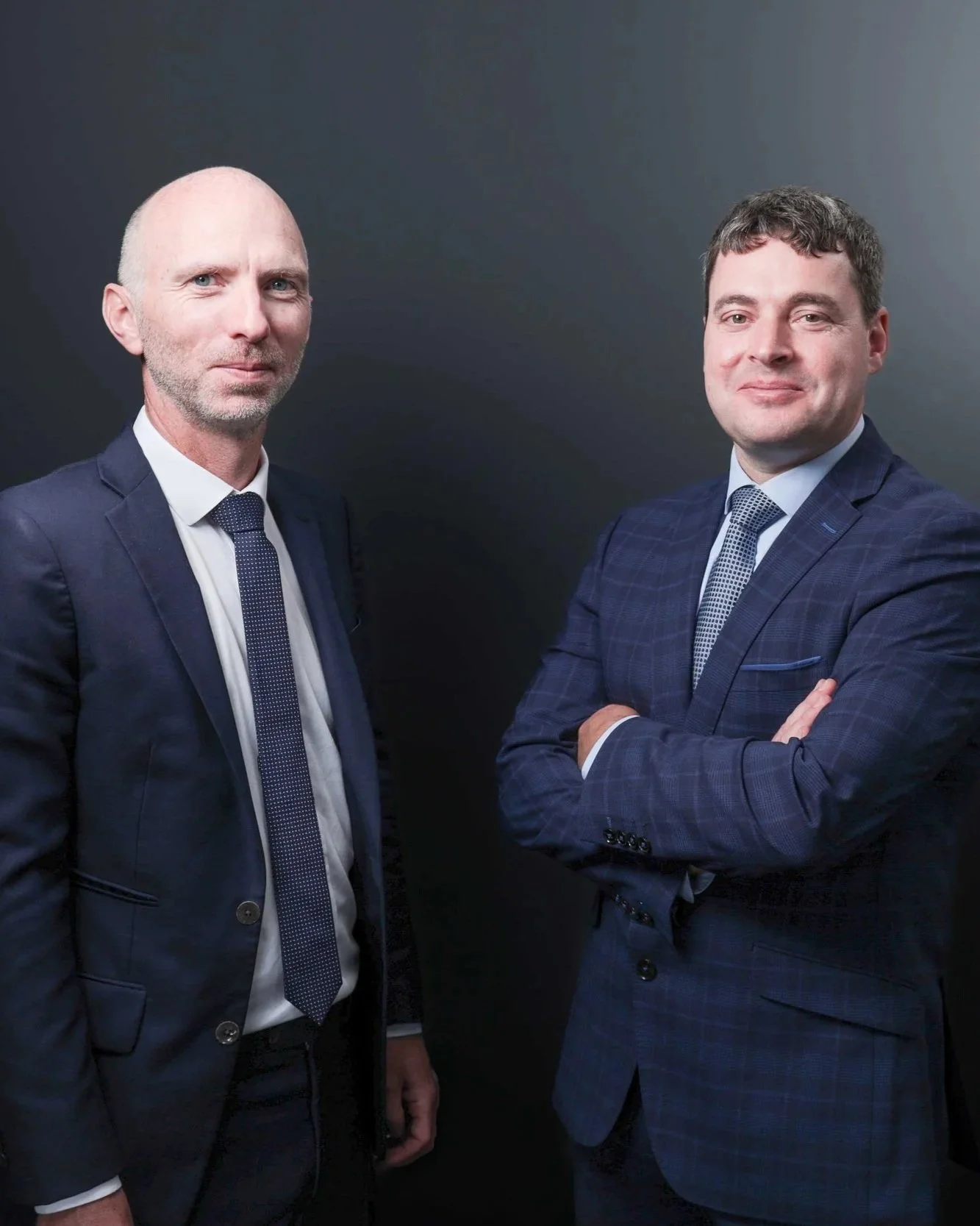 Two men in suits standing against a dark background, one with arms crossed and the other with hands at his sides.