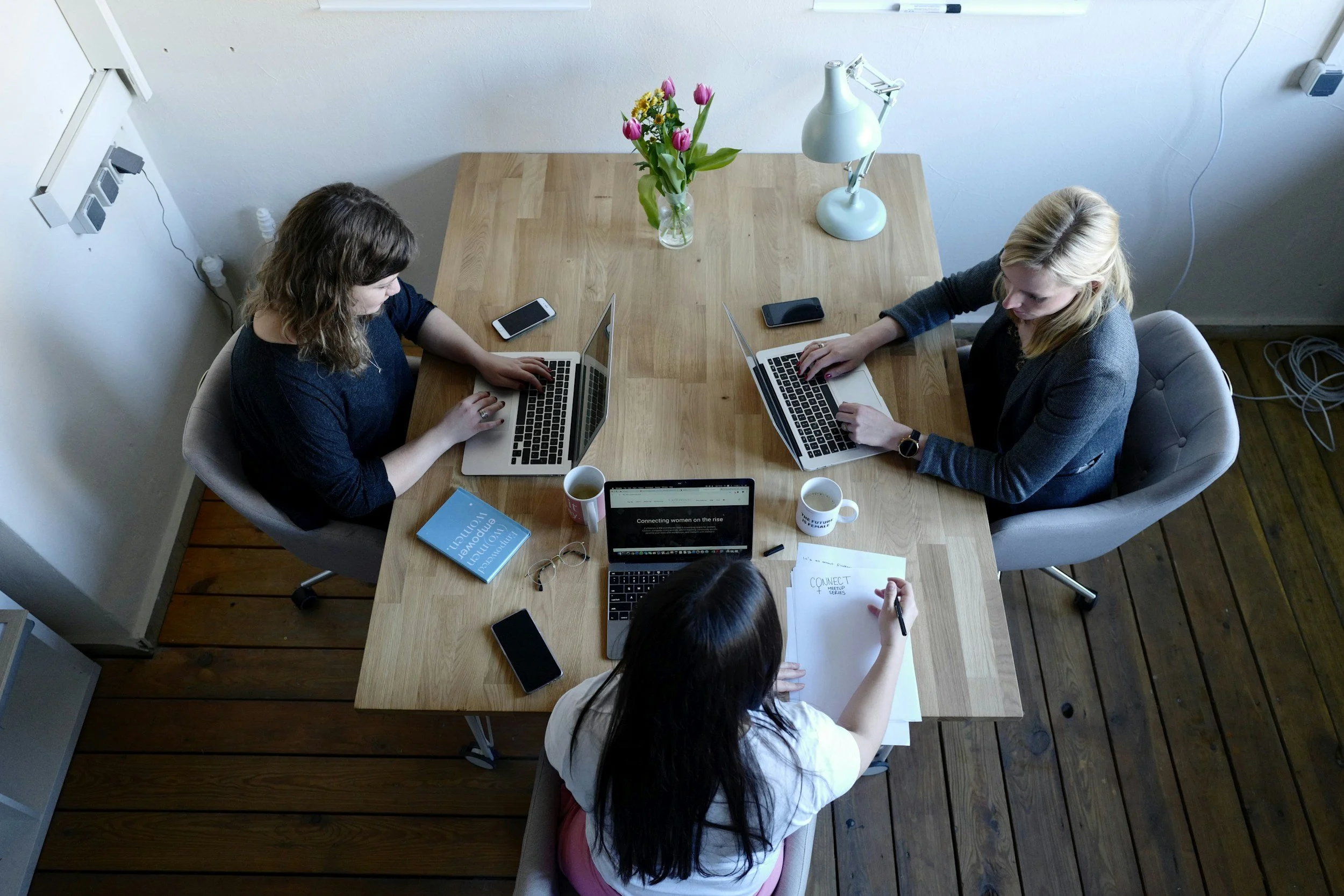 Three women working together at a wooden table with laptops, smartphones, coffee mugs, glasses, a notebook, and a pen in a bright, modern office.