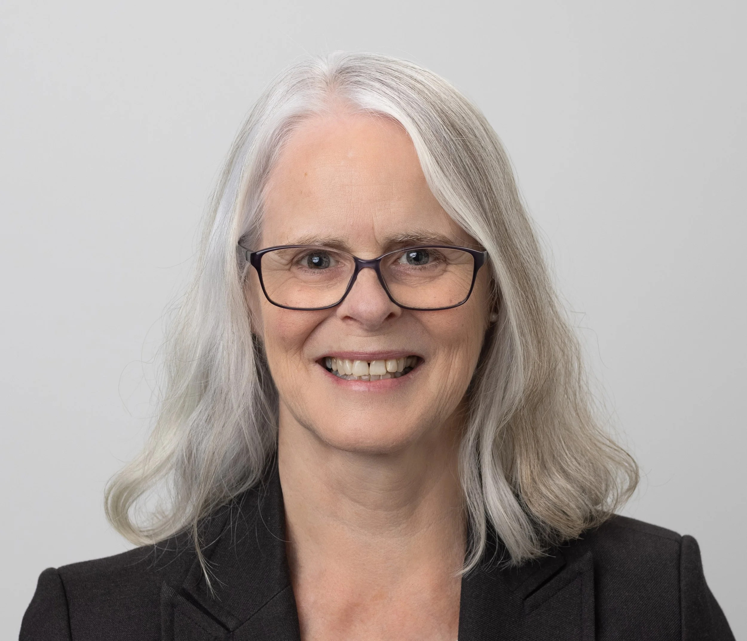 A woman with long gray hair, wearing glasses and a black blazer, smiling against a plain gray background.