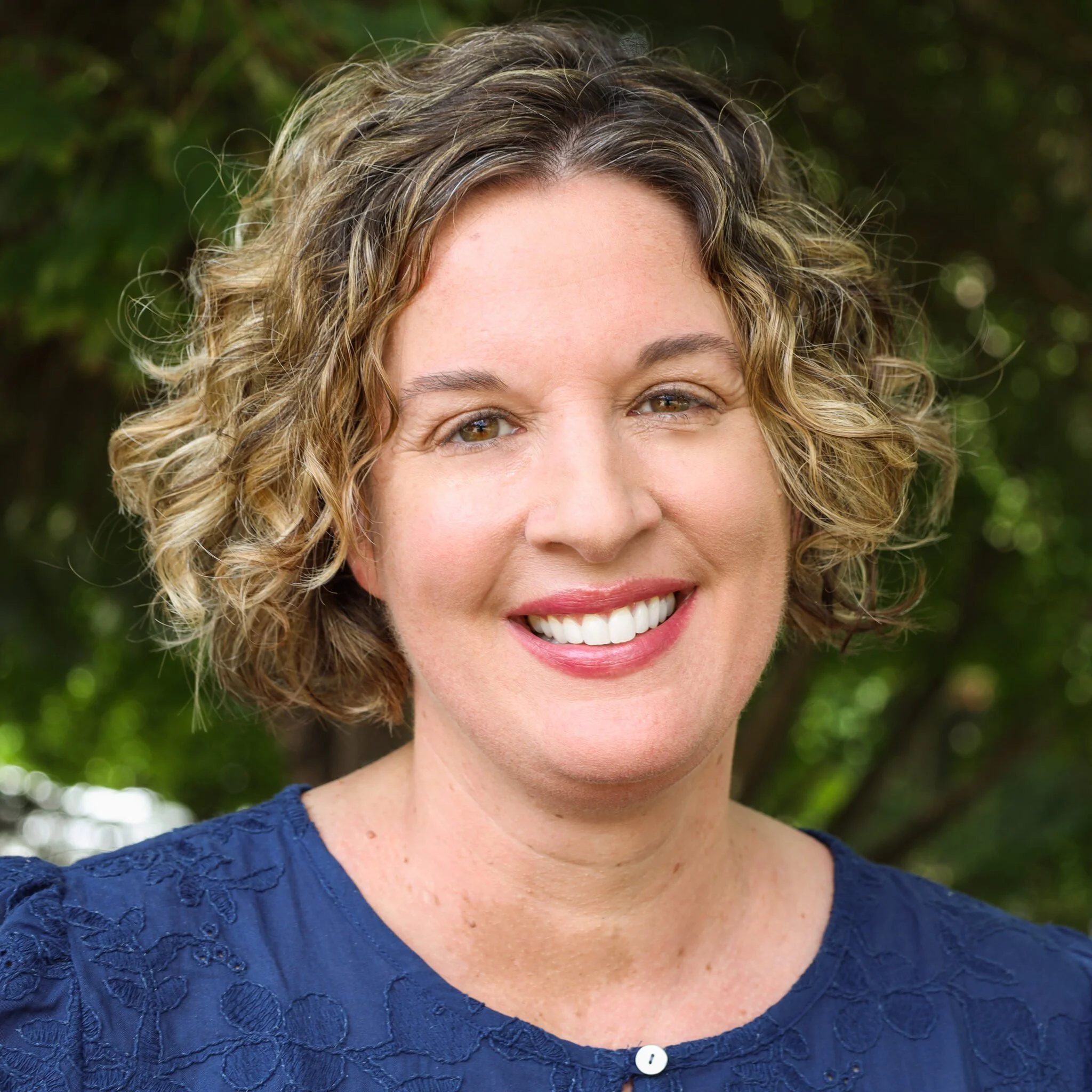 Close-up portrait of a woman with curly blonde hair, smiling, wearing a navy blue top, outdoors with green trees in the background.