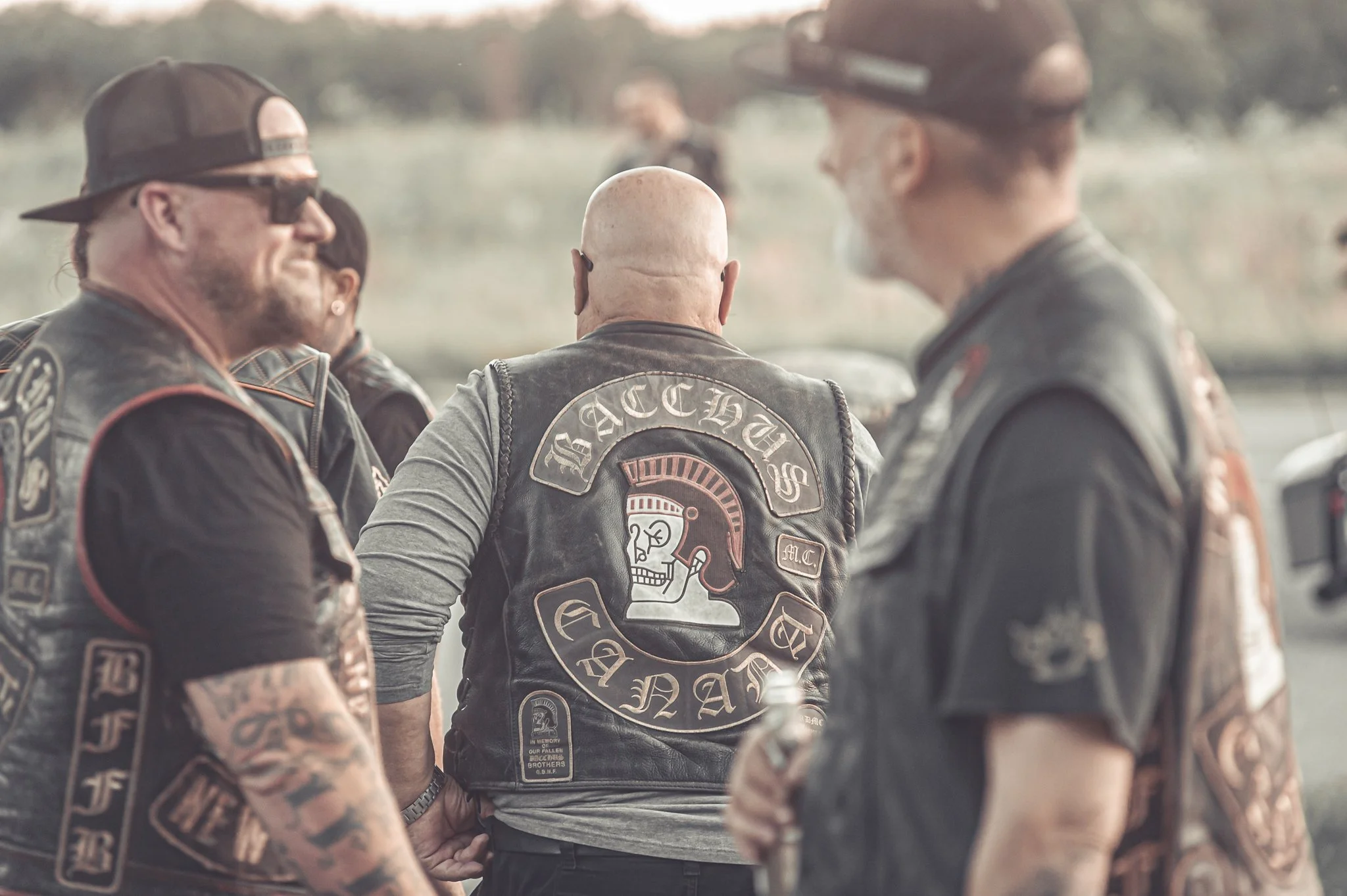 A group of bikers wearing leather vests with patches, gathered outdoors in a rural area during daytime, with a field and trees in the background.