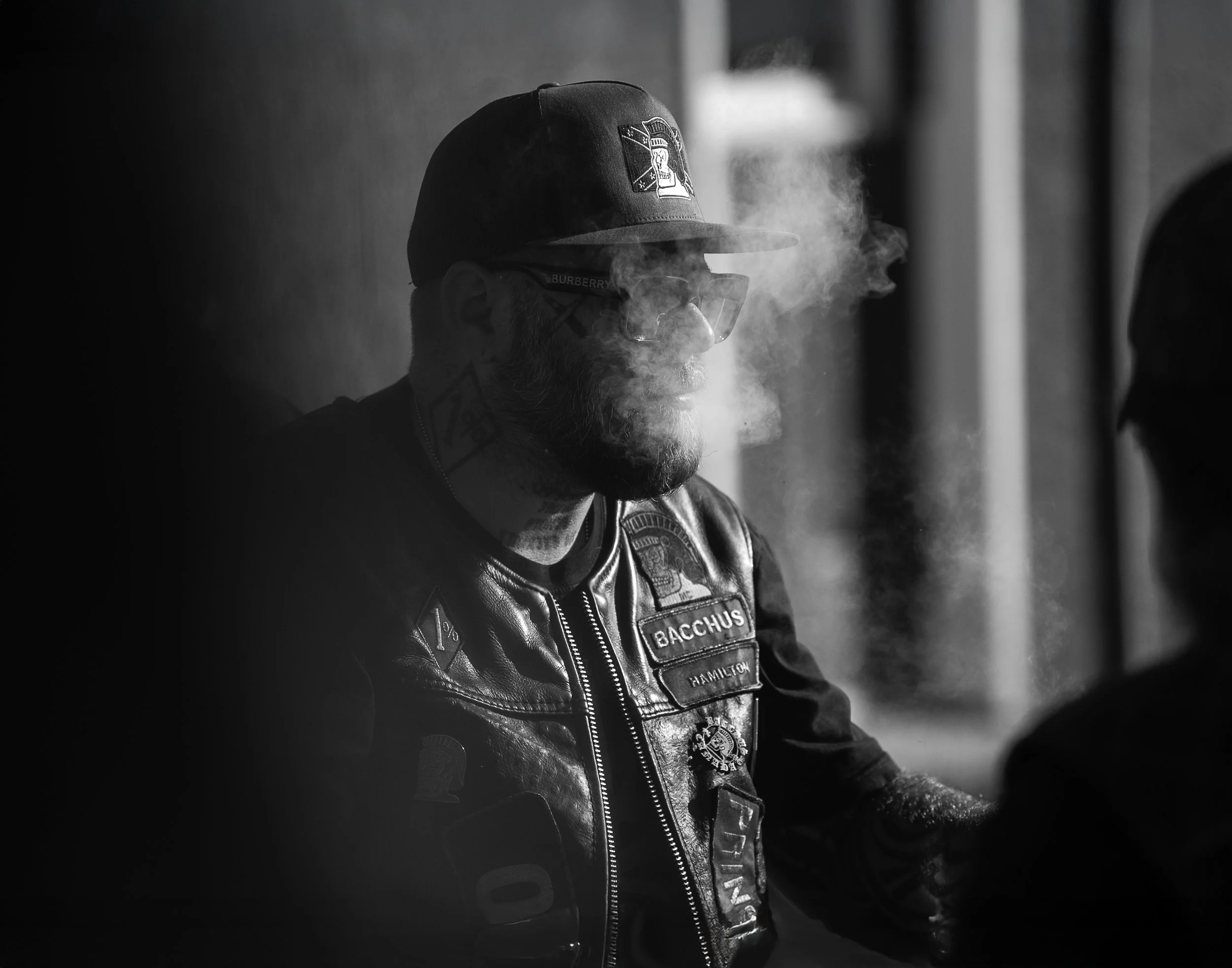 A man with a beard wearing sunglasses, a cap, and a leather vest with patches is exhaling smoke. The background is blurred.
