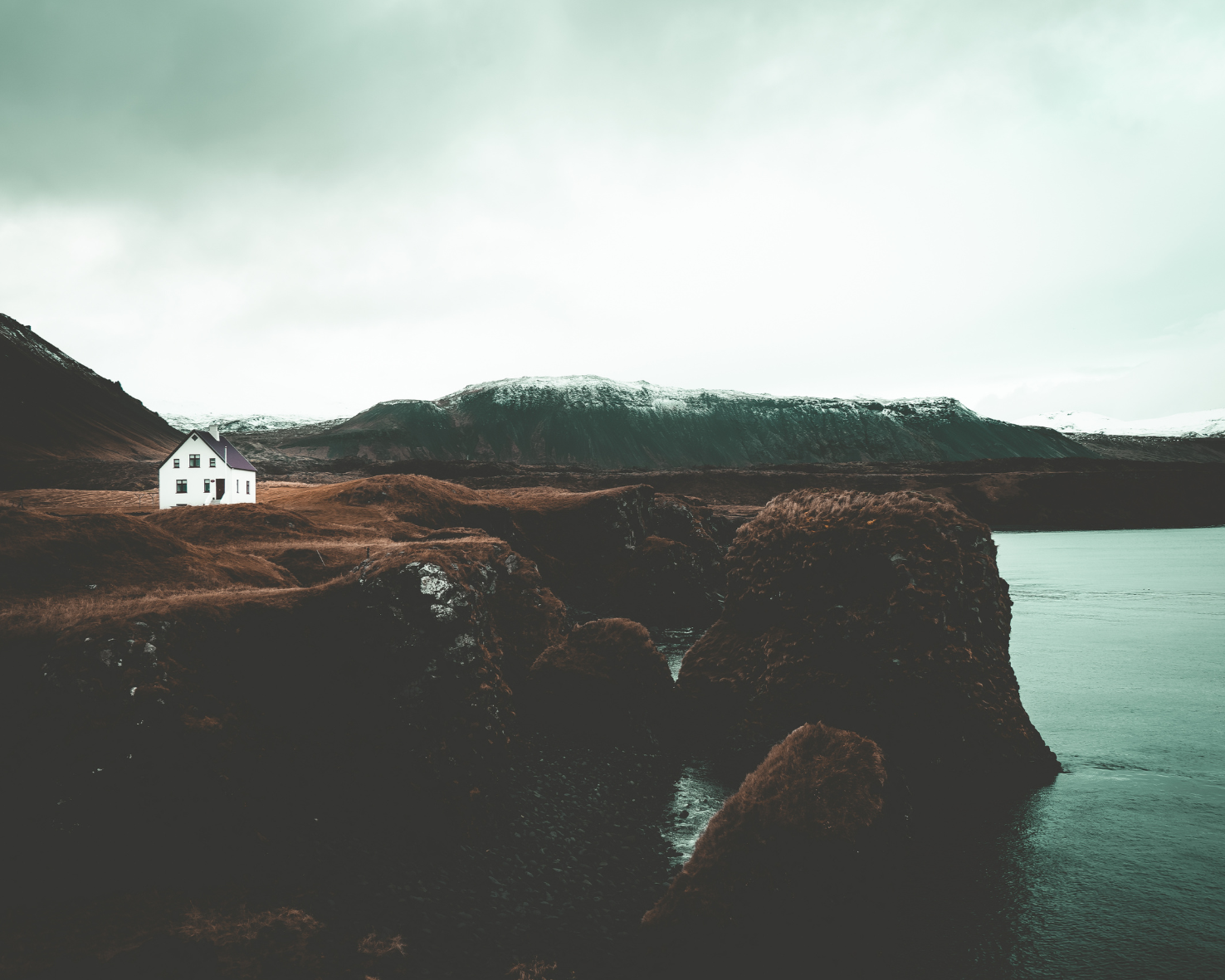A white house with a dark roof on a grassy, rocky coastline with mountains in the background and a body of water in the foreground.