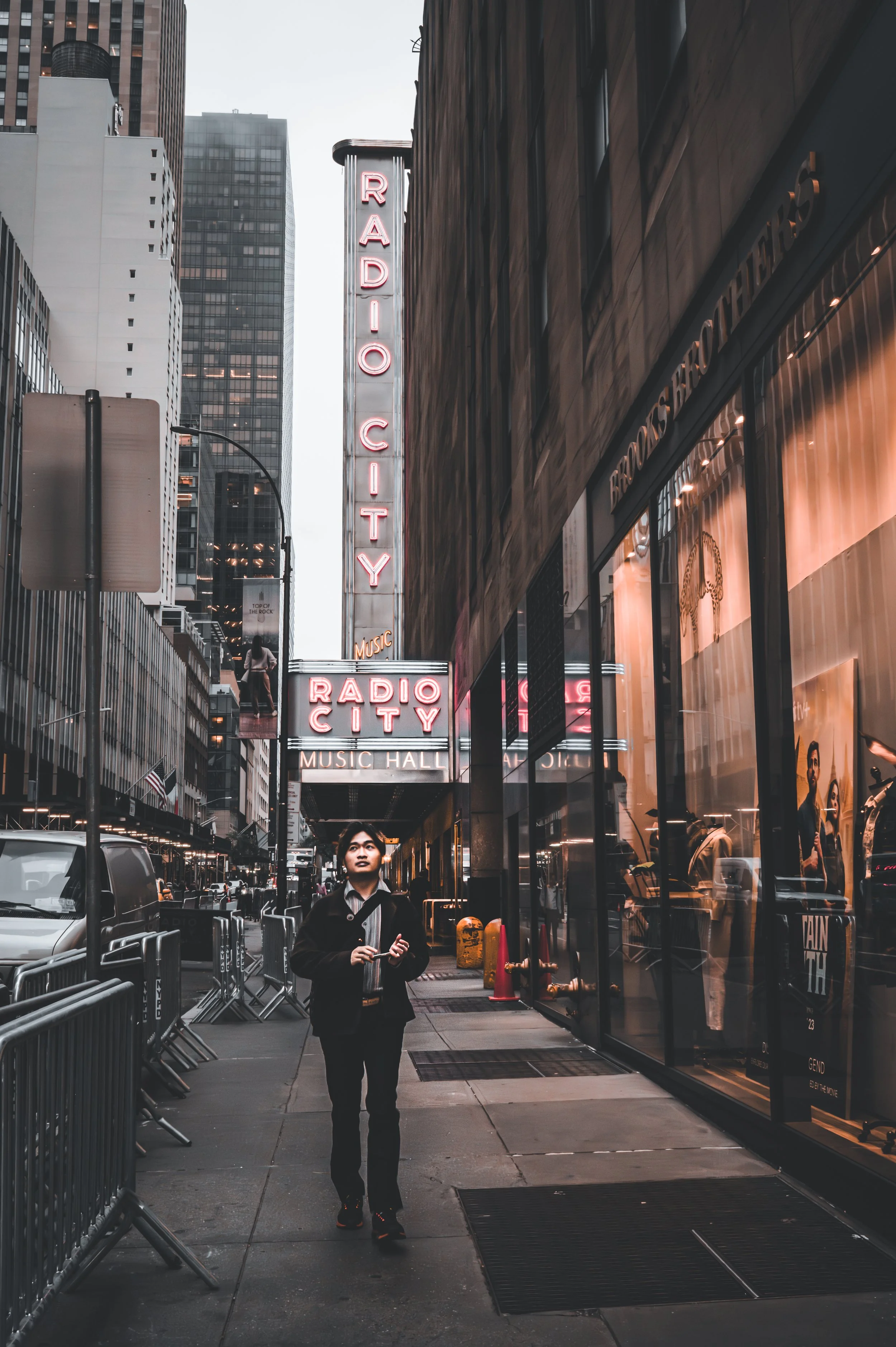 Street view showing a woman walking on a city sidewalk in front of Radio City Music Hall with its neon sign, tall skyscrapers, and storefronts on either side.