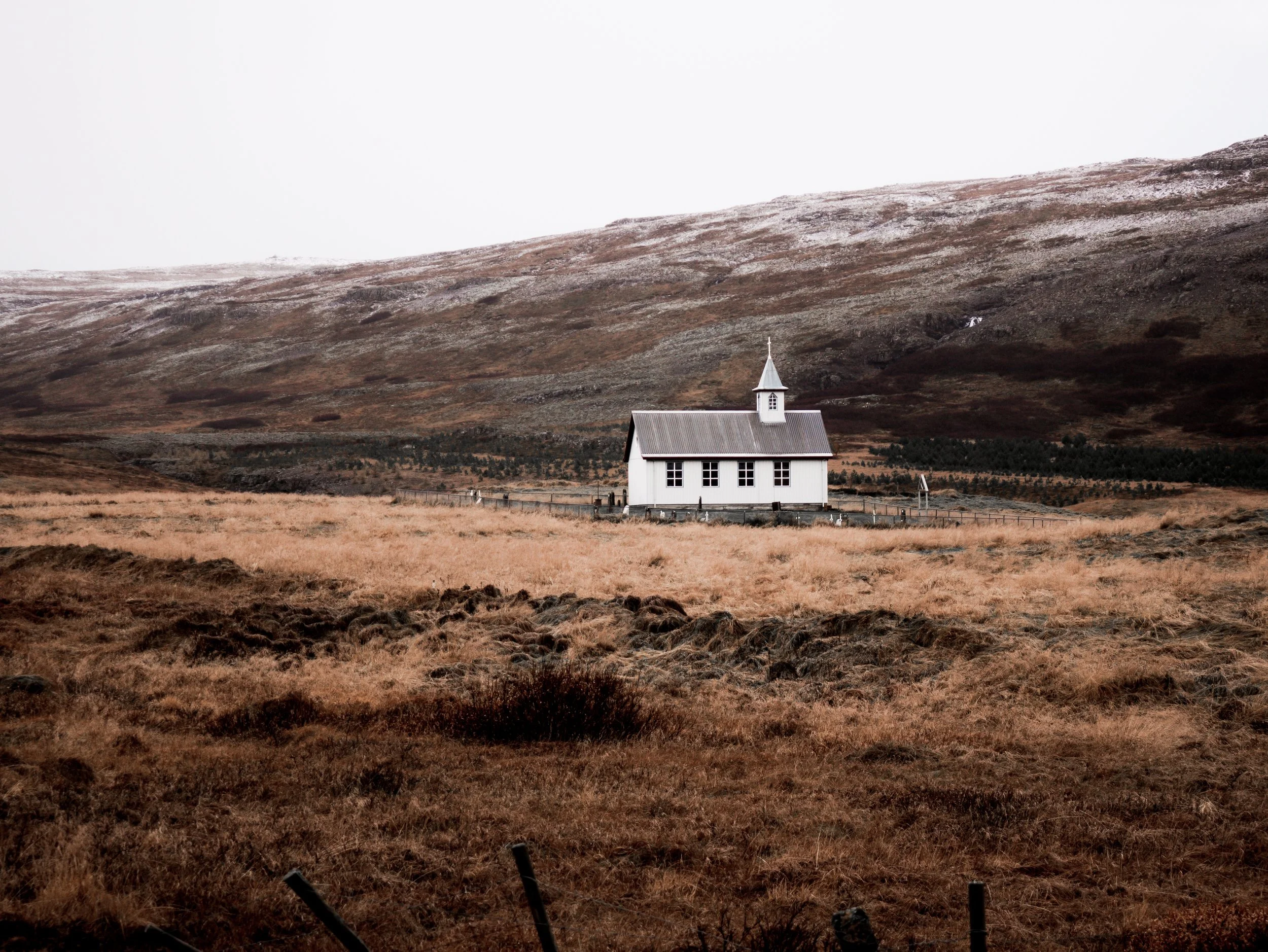 A small white church with a steeple situated in a vast, open landscape with brown grass and distant mountains under a cloudy sky. Iceland 