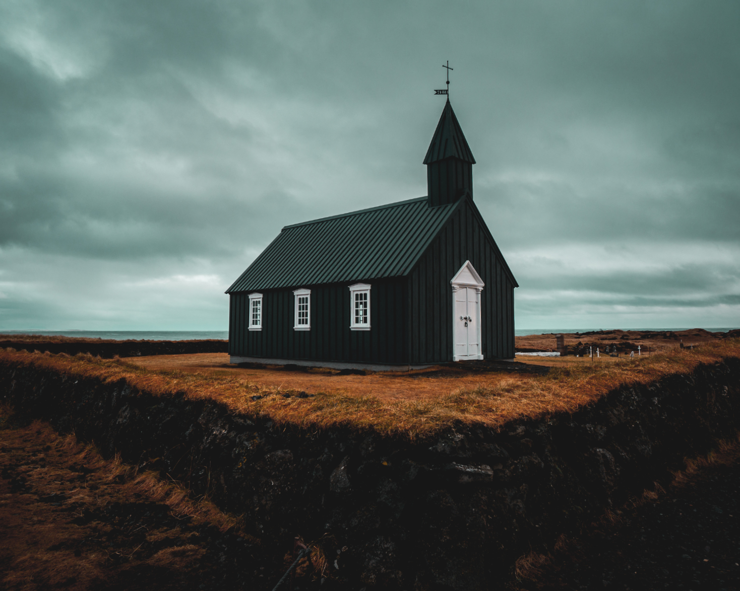 A small black wooden church in Iceland with white trim on a grassy landscape under a cloudy sky.