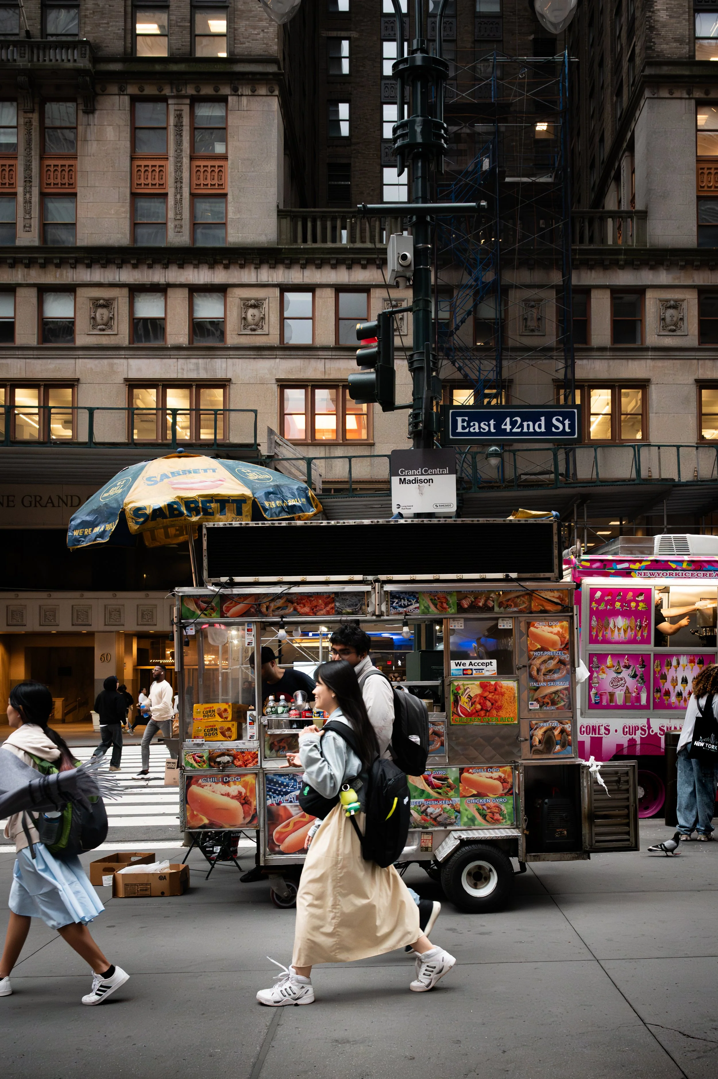 Street scene in New York City showing a food cart with people walking by on East 42nd Street near Grand Central Madison, with buildings and street signs visible.