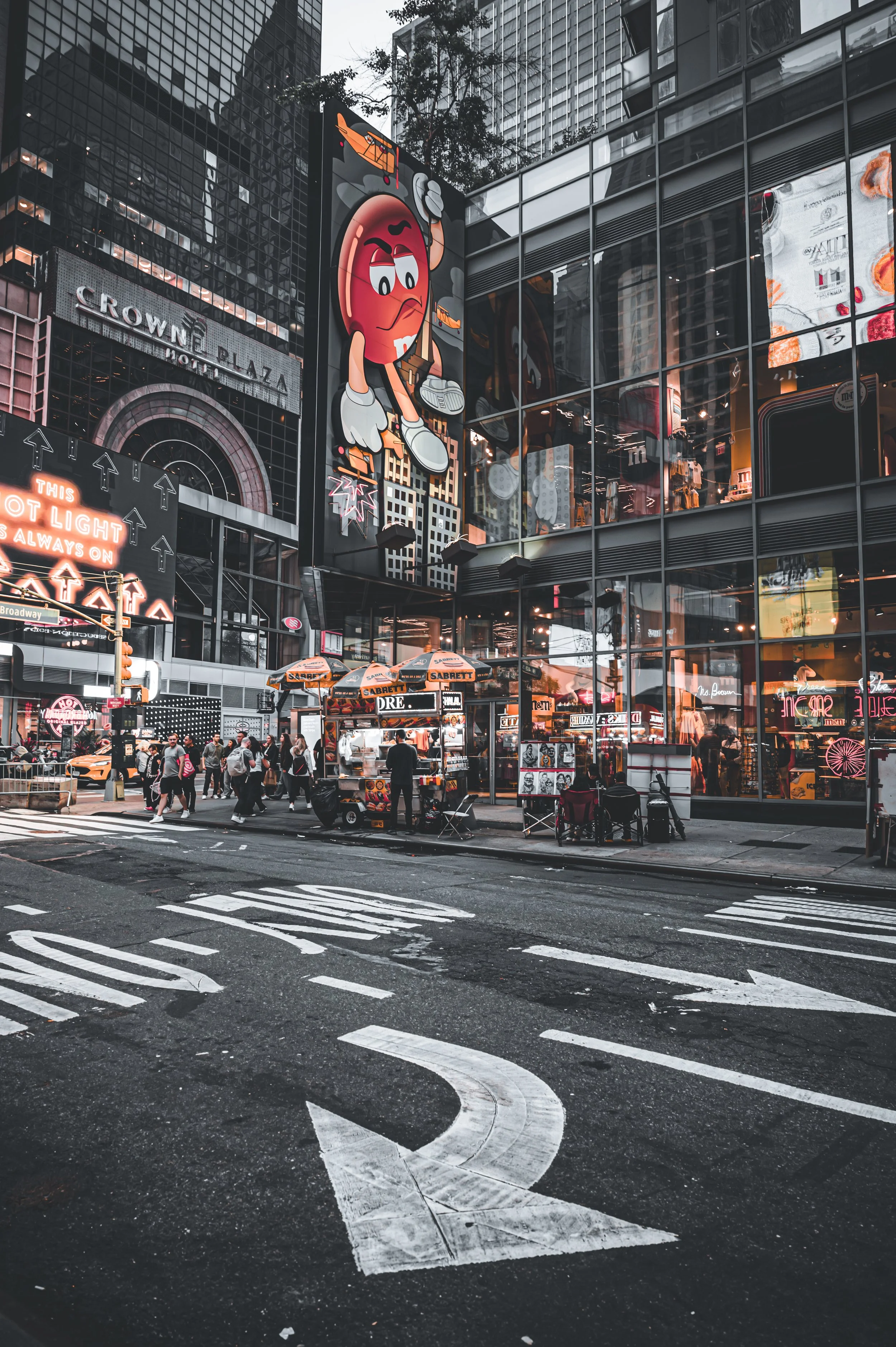 A busy city street scene at dusk with pedestrians crossing and sitting outside. There are food carts and bright neon signs, including a large animated cartoon character on a building facade. The scene features modern glass buildings and a mix of comm