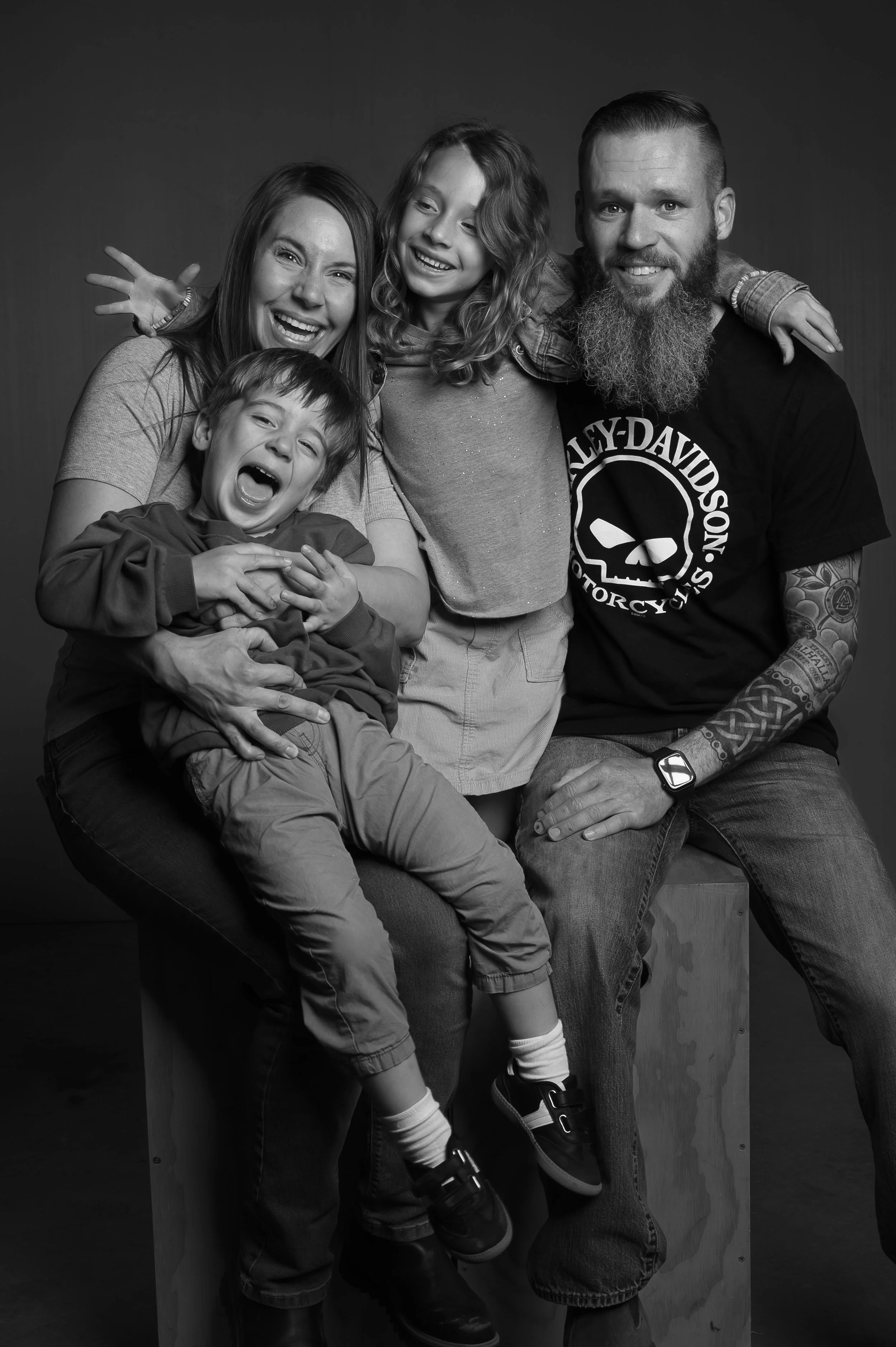 Black and white photo of a family of five, smiling and having fun together, with the father wearing a Harley Davidson t-shirt, the mother with her arm around the daughter, a young boy sitting on the mother's lap, and a young girl with curly hair lean