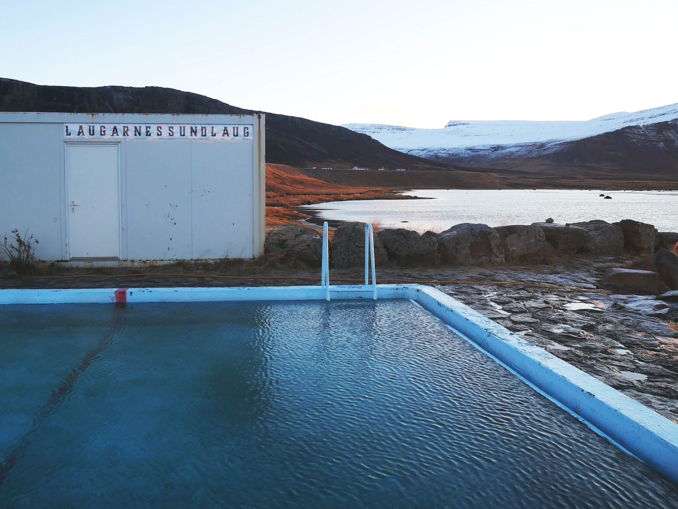 An outdoor swimming pool with metal handrails, set in a rugged landscape with a lake, mountains, and snow in the distance, under clear skies. Iceland hot springs.