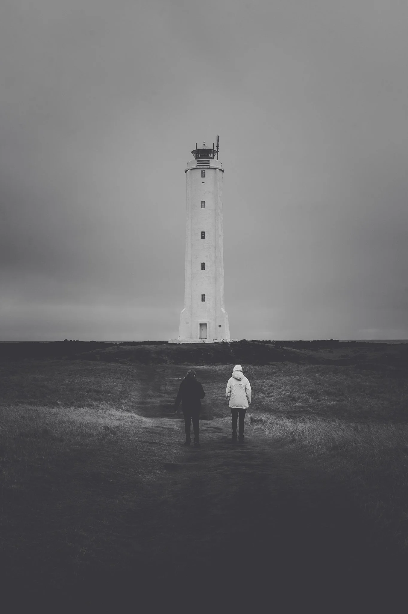 Two people walking on a path toward a lighthouse on a cloudy day in Iceland.