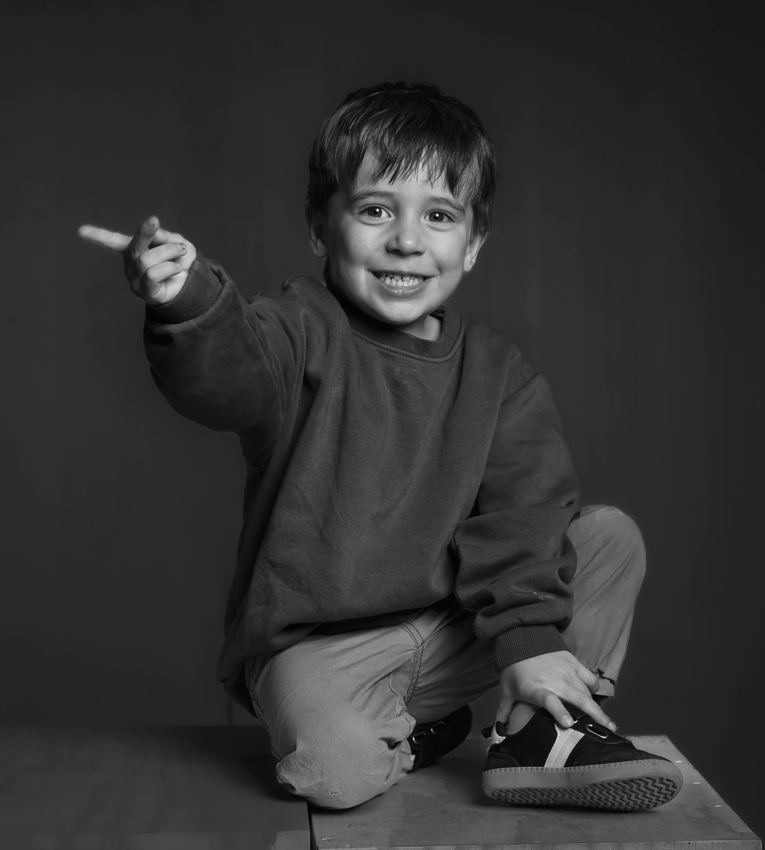 Black and white photo of a smiling young boy crouching down, pointing to his left, wearing a hoodie, jeans, and sneakers, with a dark background.