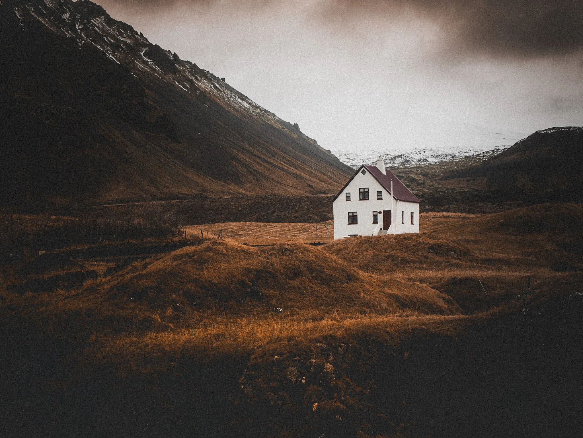 A white house with a red roof sits in a desolate landscape of rolling brown hills and mountains under a cloudy sky. Iceland